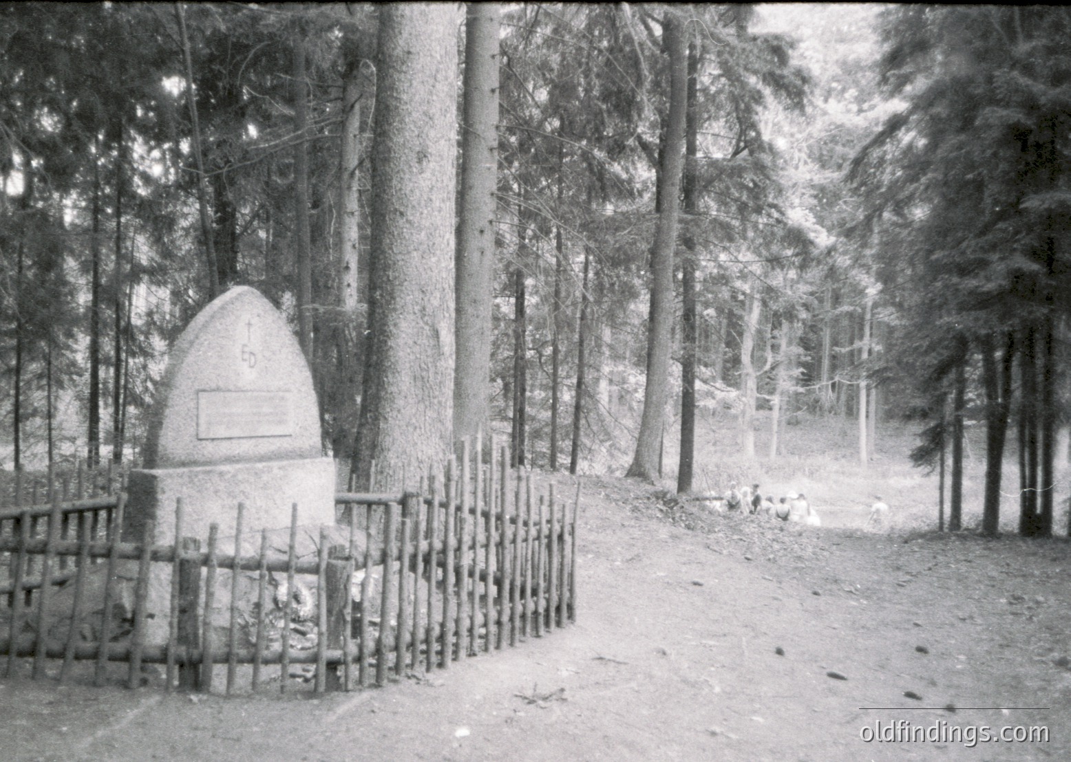 Monumental stone obelisk with engraved plaque in a wooded forest path, surrounded by a rustic wooden fence. Snow covers ground and branches, suggesting winter. Likely a memorial or historical marker.