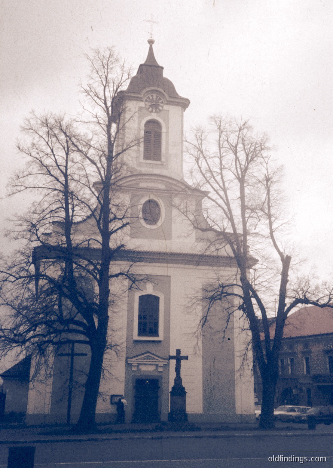 Neoclassical church with tall bell tower featuring a clock and cross, flanked by bare trees. Architectural details include arched windows and a symmetrical façade. Likely Eastern European, late 19th to early 20th century.