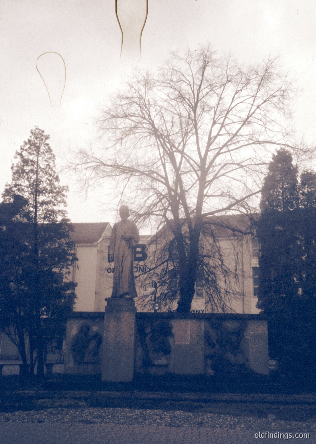 Statue of a robed figure on pedestal in courtyard, flanked by trees and a building with Cyrillic lettering. Likely Eastern European institutional or academic setting, mid-20th century.