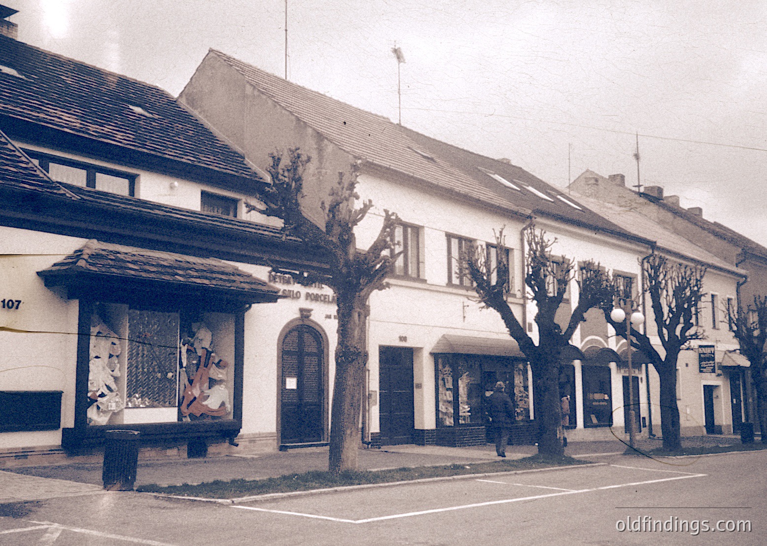Mid-20th century European street corner with two-story brick buildings. Left: "Pizzeria" sign with vintage mural of figures in window. Right: "Hotel" sign with arched entrance. Bare trees and minimalist street lamps. Black-and-white, likely 1950s-1960s.