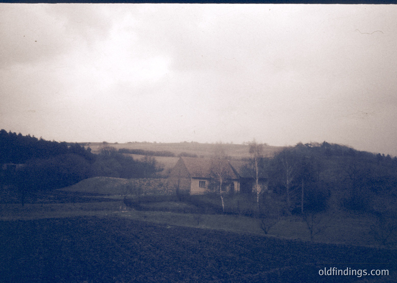Vintage sepia-toned rural scene featuring a single-story farmhouse with a pitched roof, surrounded by open fields and leafless trees. The landscape includes rolling hills and sparse vegetation, suggesting a cold season. Likely Eastern European countryside, mid-20th century.