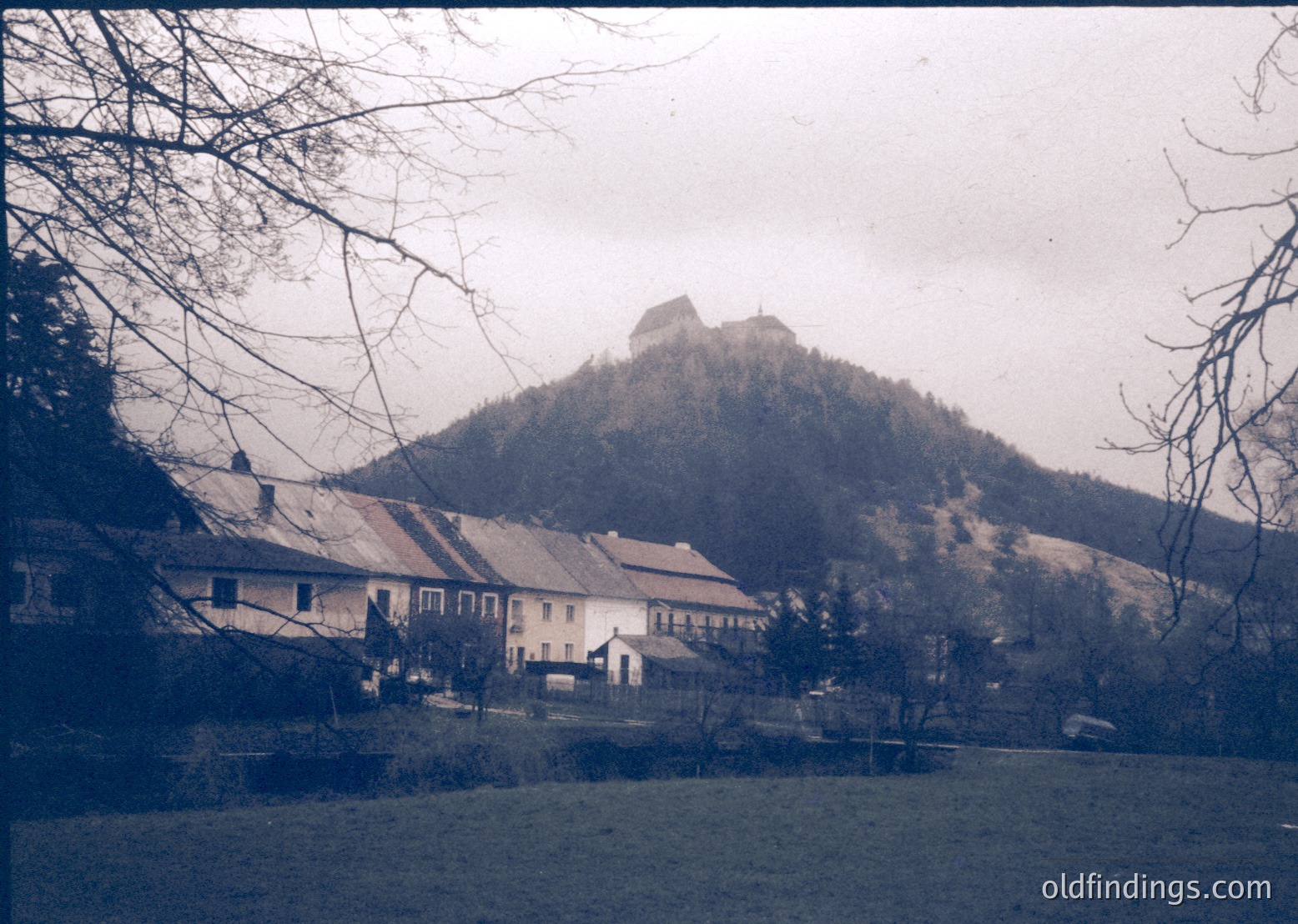 Vintage black-and-white village scene with clustered brick houses under a rocky hilltop fortress. Bare winter trees frame the composition. Likely Eastern European rural architecture, mid-20th century.