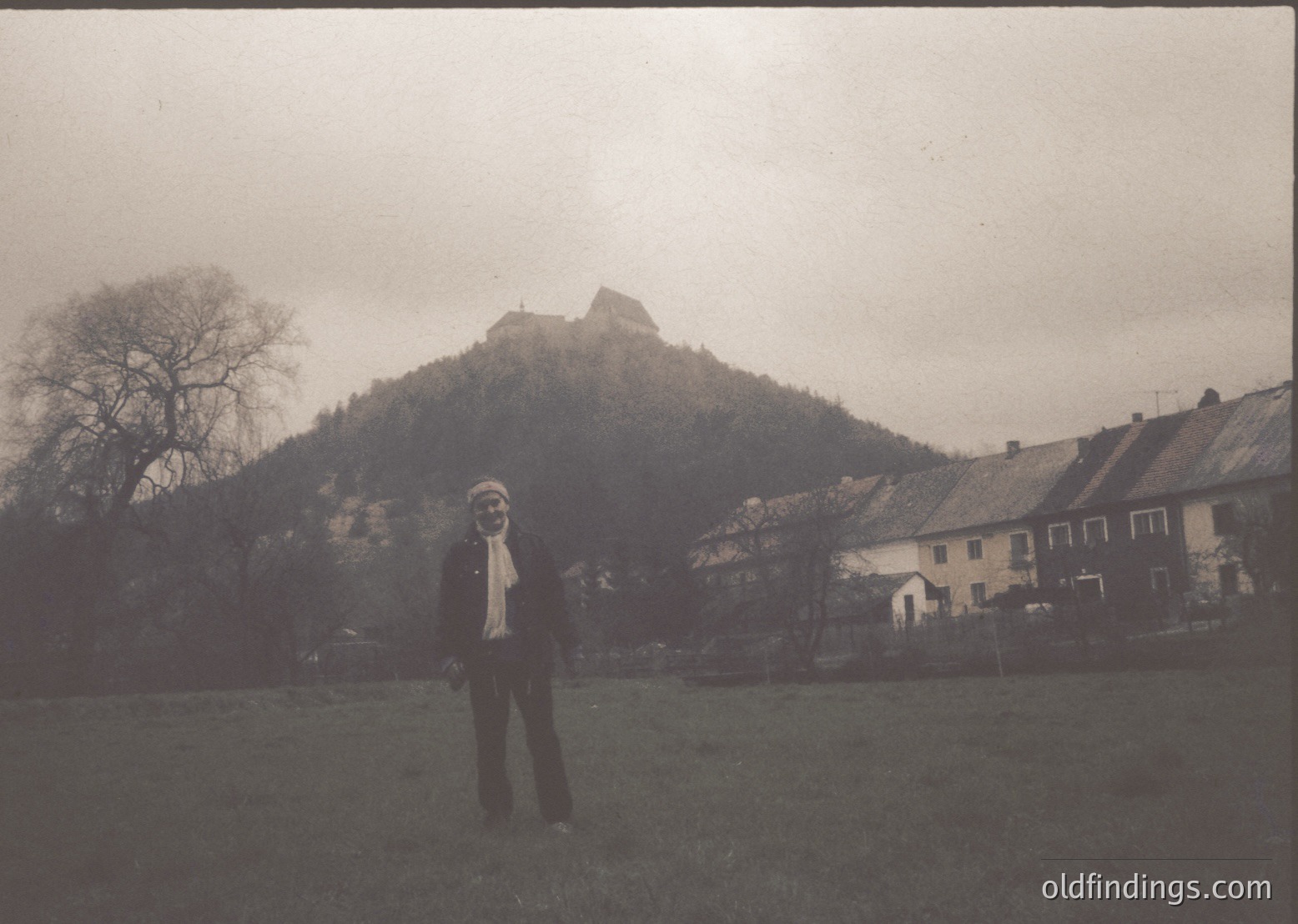 Vintage black-and-white photo of a man in 1970s-era attire—wide-lapelled jacket, scarf, and trousers—posing in a rural European village. A hilltop castle or fortress looms in the background, surrounded by modest stone houses with sloped roofs. Overcast sky enhances the nostalgic atmosphere. Likely or due to architectural style.