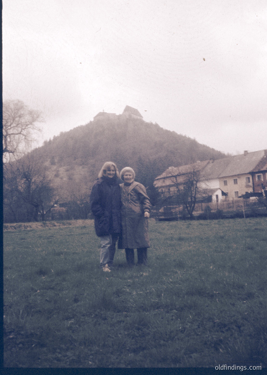 Vintage black-and-white photo of two women posing in a grassy field with a rocky hillock and rustic houses in background. Mid-20th century European countryside setting, likely 1950s–1960s. Warm winter coats and scarves suggest chilly weather.