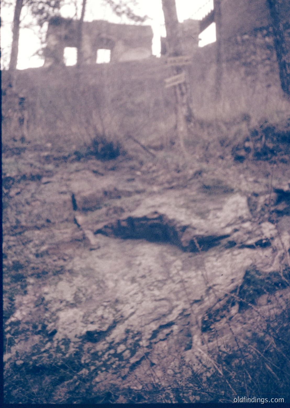 Vintage sepia-toned photo of a rural, abandoned stone structure surrounded by overgrown vegetation. Crumbling walls and a collapsed roof suggest neglect, with a wooden signpost partially visible. Likely Eastern European countryside, mid-20th century.