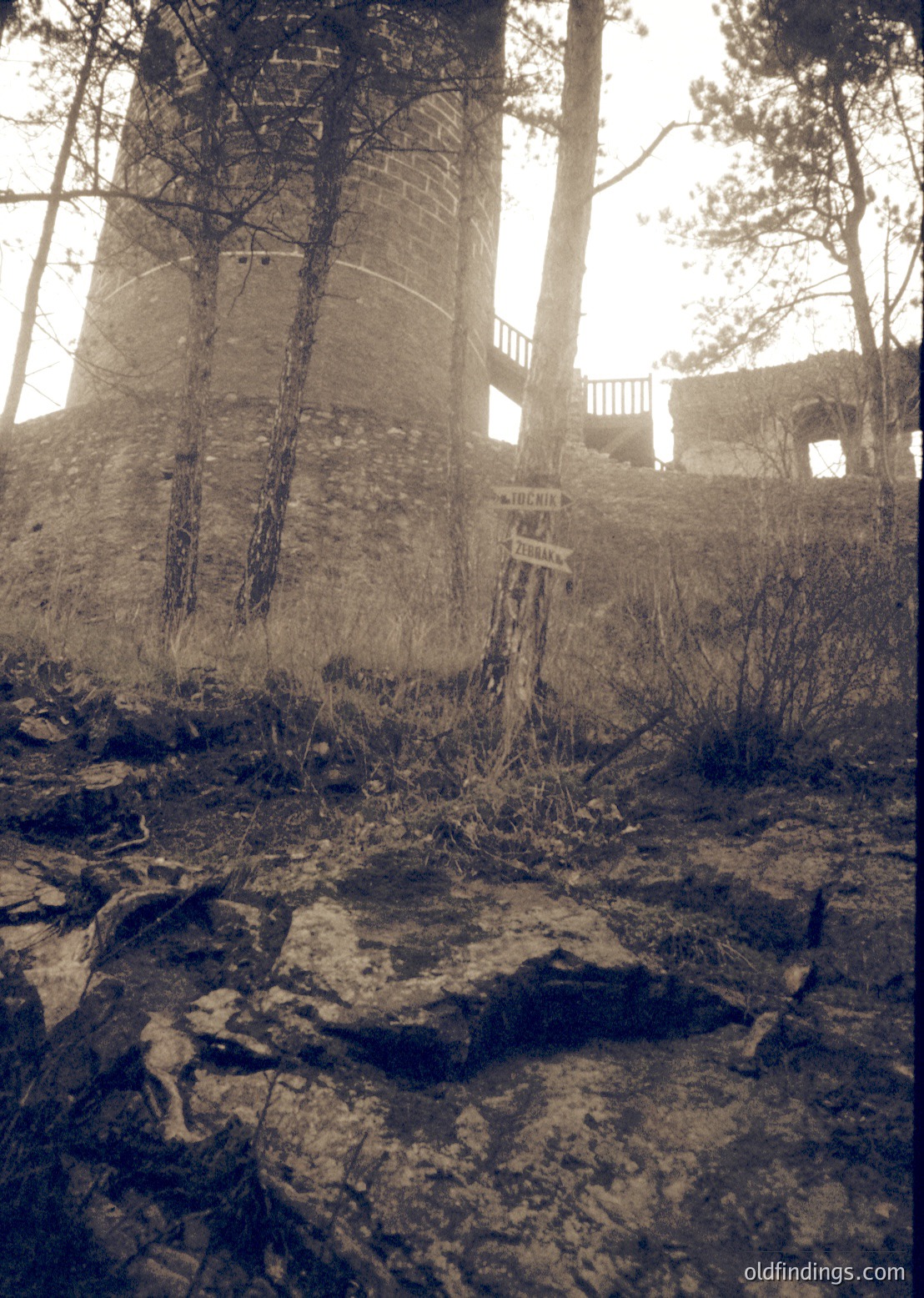A sepia-toned photograph of a weathered, abandoned stone structure with a sign reading "TOCHIN" and "ПЕРЕСЕЛЕНИЕ" (meaning "resettlement" in Bulgarian). The ruins include a spiral staircase and remnants of a balcony, surrounded by overgrown vegetation and rocky terrain. Likely from a Soviet-era mining or industrial site in Bulgaria, dated mid-20th century.