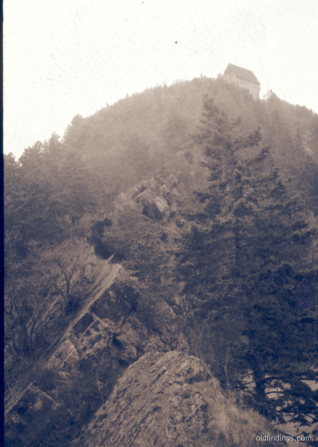Vintage sepia-toned photograph of a mist-shrouded hilltop fortress atop rugged terrain, likely Eastern European. Stone tower and walls perched on rocky outcrop, surrounded by dense forest. Path winding up the slope suggests historical or medieval architecture.