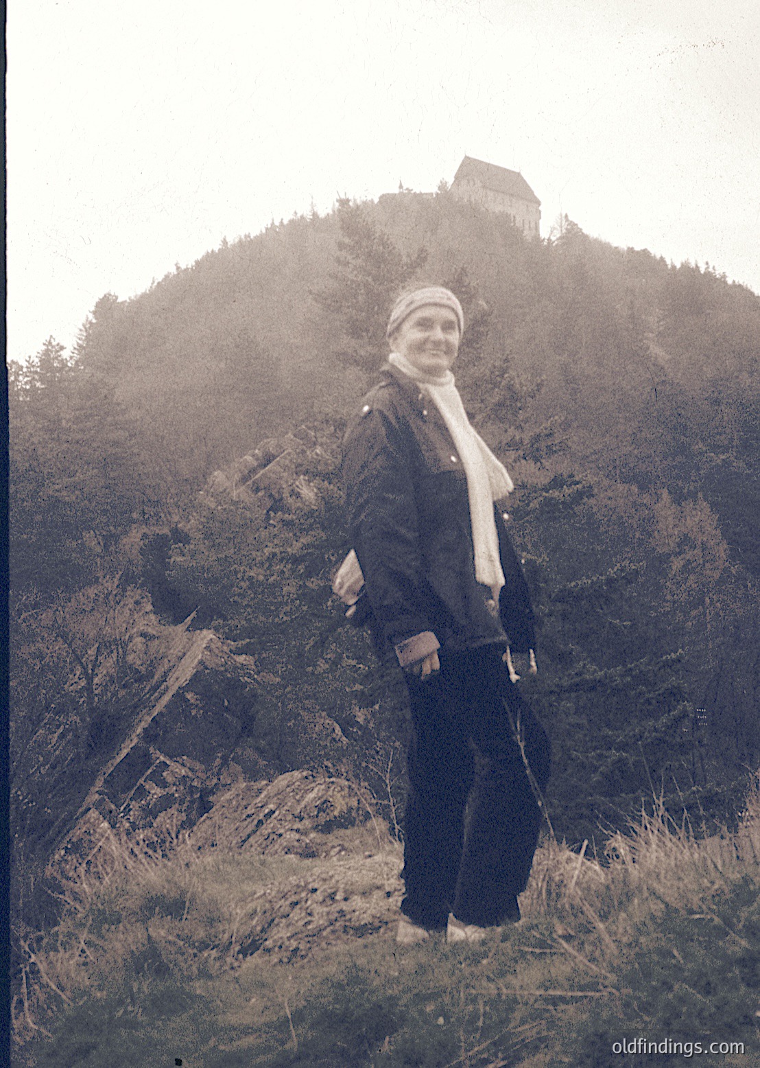 Vintage sepia-toned portrait of a woman in outdoor attire—dark jacket, patterned scarf, and trousers—standing on a rocky hillside near a medieval tower. The scene suggests a European alpine or hilly region, likely 1950s–1960s.