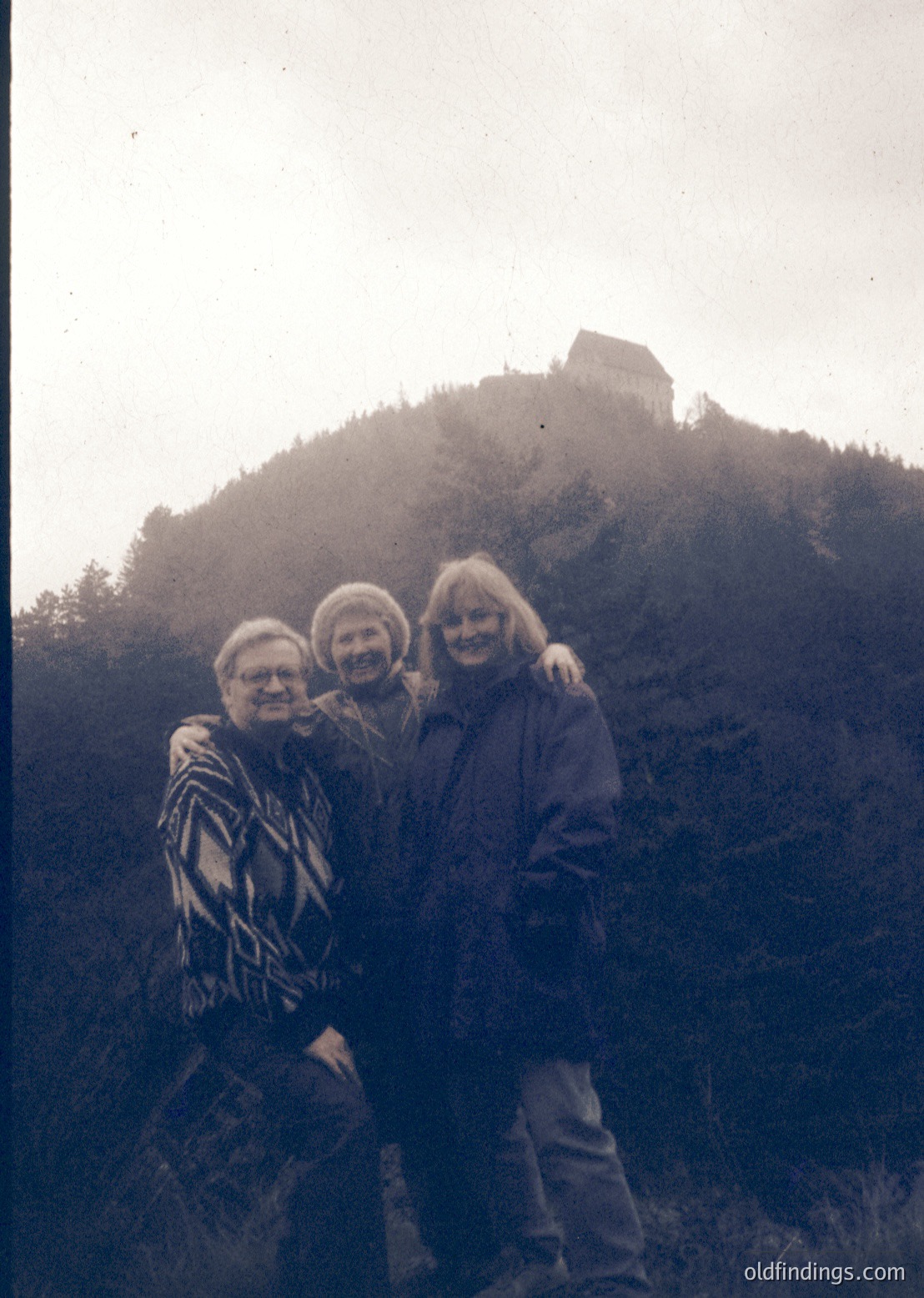 Vintage black-and-white photo of three adults posing on a misty hillside, with a medieval fortress ruin in background. Mid-20th century clothing suggests or . Natural lighting enhances atmospheric mood.