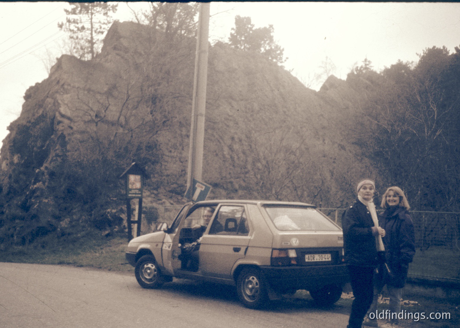 Vintage car parked beside rugged rock formations with two women in 1970s-style clothing. The car’s license plate suggests Eastern European origin. Scenic roadside setting with natural rock backdrop.