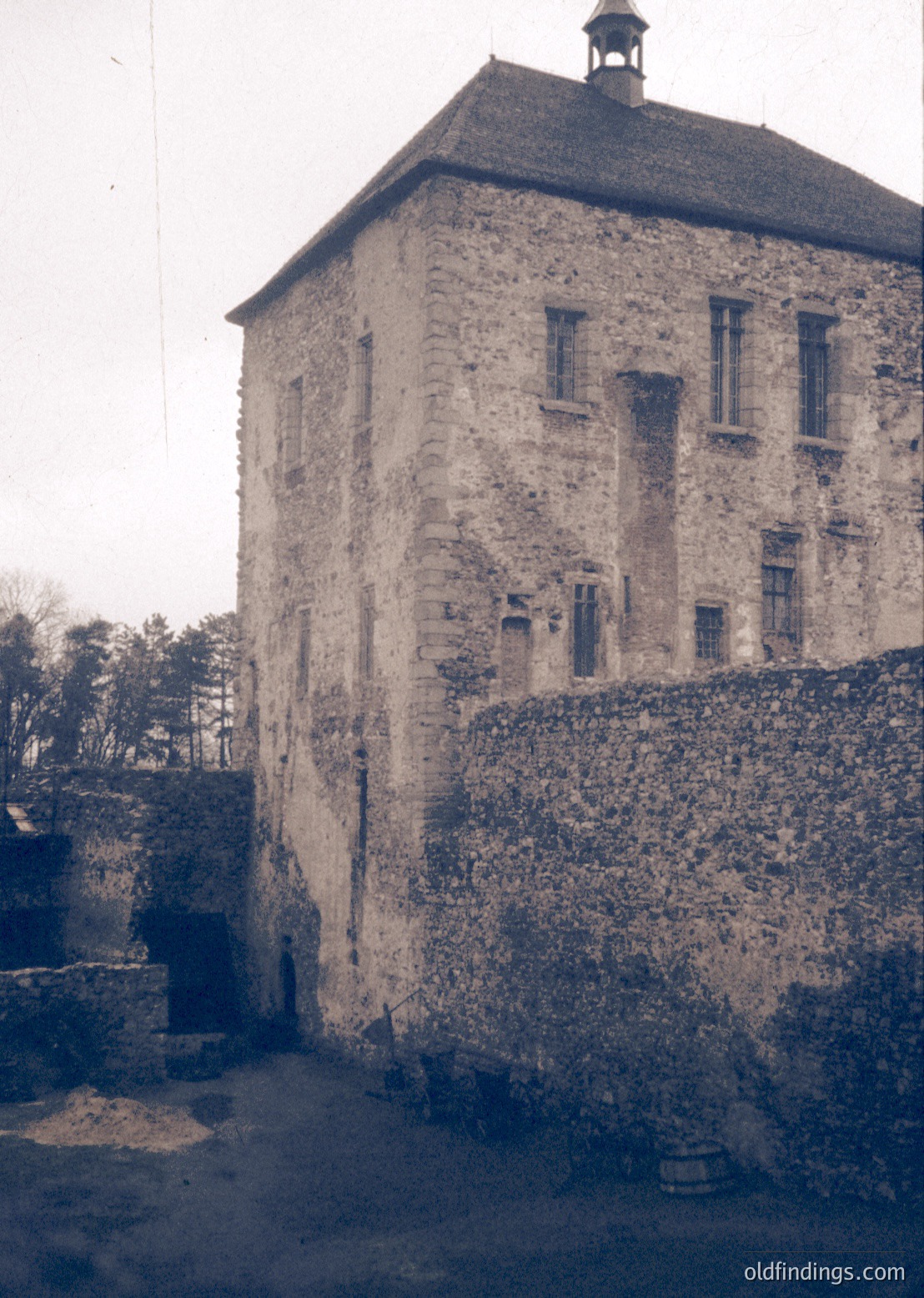 Three-story stone fortress tower with crenellated roofline, flanked by high defensive walls. Small rectangular windows and a small turret atop. Foreground shows weathered stone steps and a barrel in the courtyard. Likely Eastern European medieval architecture.