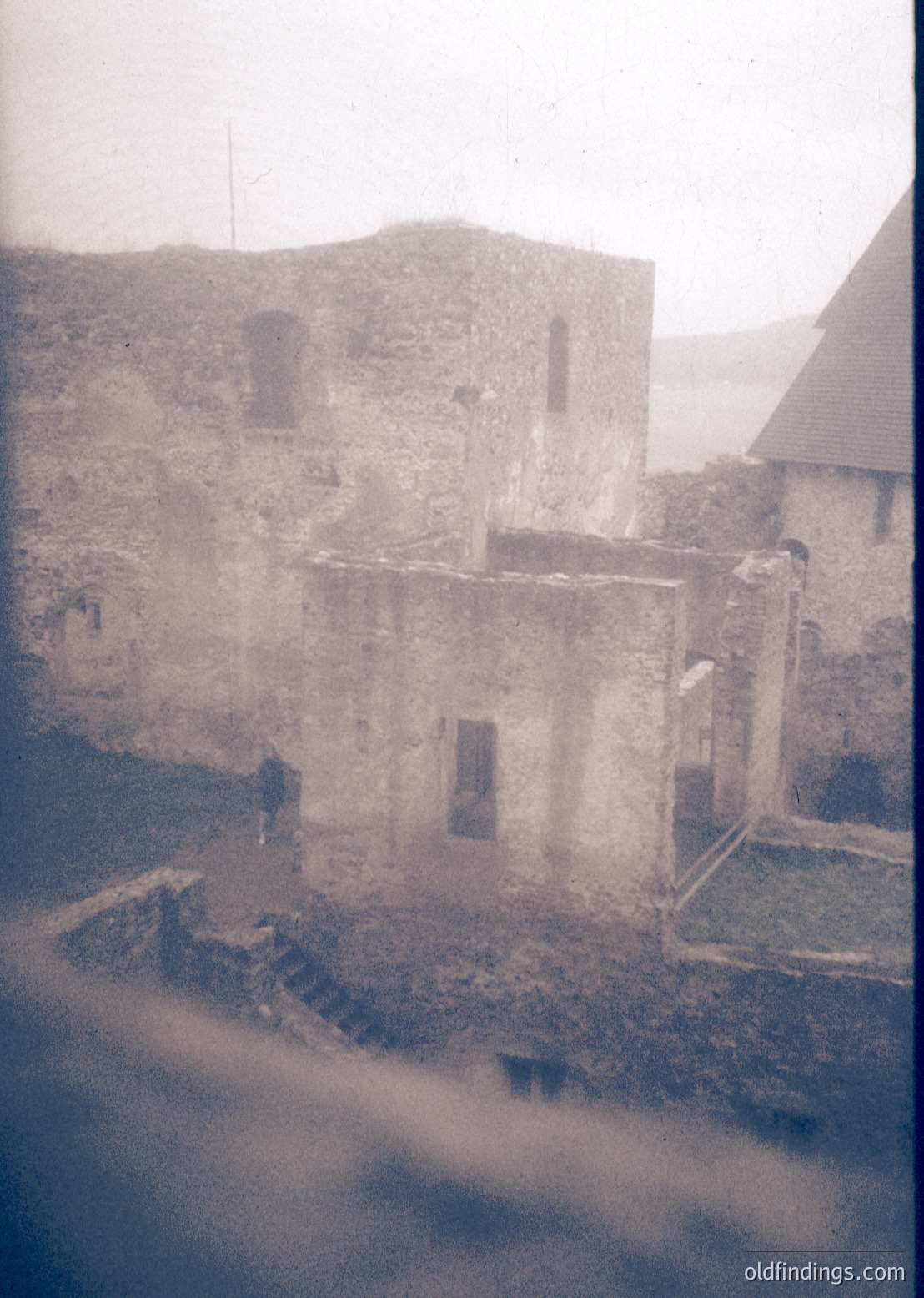 Mid-20th century black-and-white photo of weathered stone ruins with crumbling walls and arched windows. A lone figure stands on a stone staircase near a partially collapsed structure. Fog or mist obscures distant rooftops, suggesting a coastal or high-altitude setting.