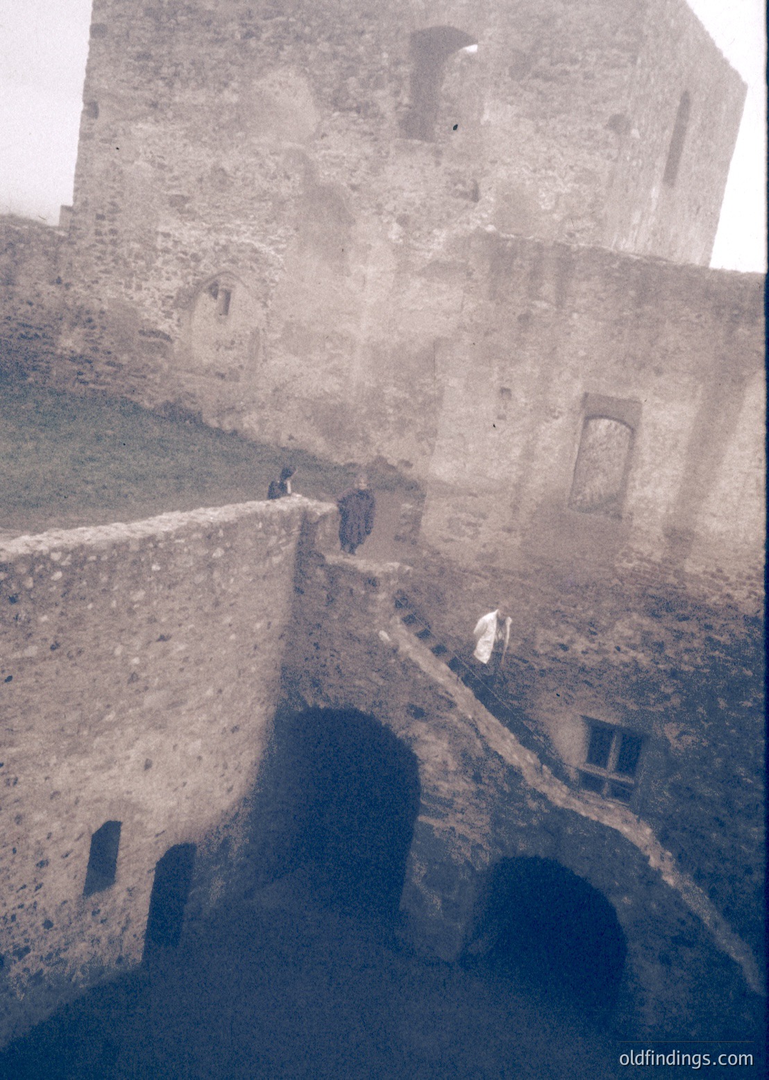 Historic stone fortress wall with arched openings and narrow pathways. Two figures in dark clothing explore the elevated ruins, suggesting a 19th–early 20th century European setting. Weathered texture and architectural details hint at military or defensive use.