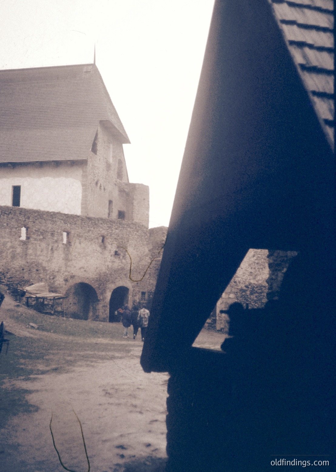 Medieval stone fortress courtyard with arched gatehouse and tower, framed by wooden beams. Likely Eastern European, 19th–early 20th century. Minimal modern elements suggest preserved historical architecture.