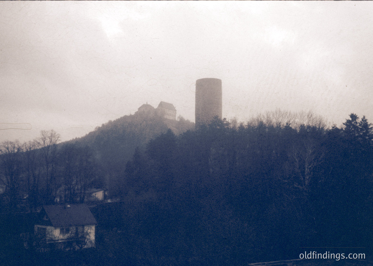 Vintage sepia-toned photo of a medieval hilltop fortress with a cylindrical tower, surrounded by sparse winter trees. Residential buildings at base suggest nearby settlement. Likely Eastern European, 20th-century industrial-era architecture.