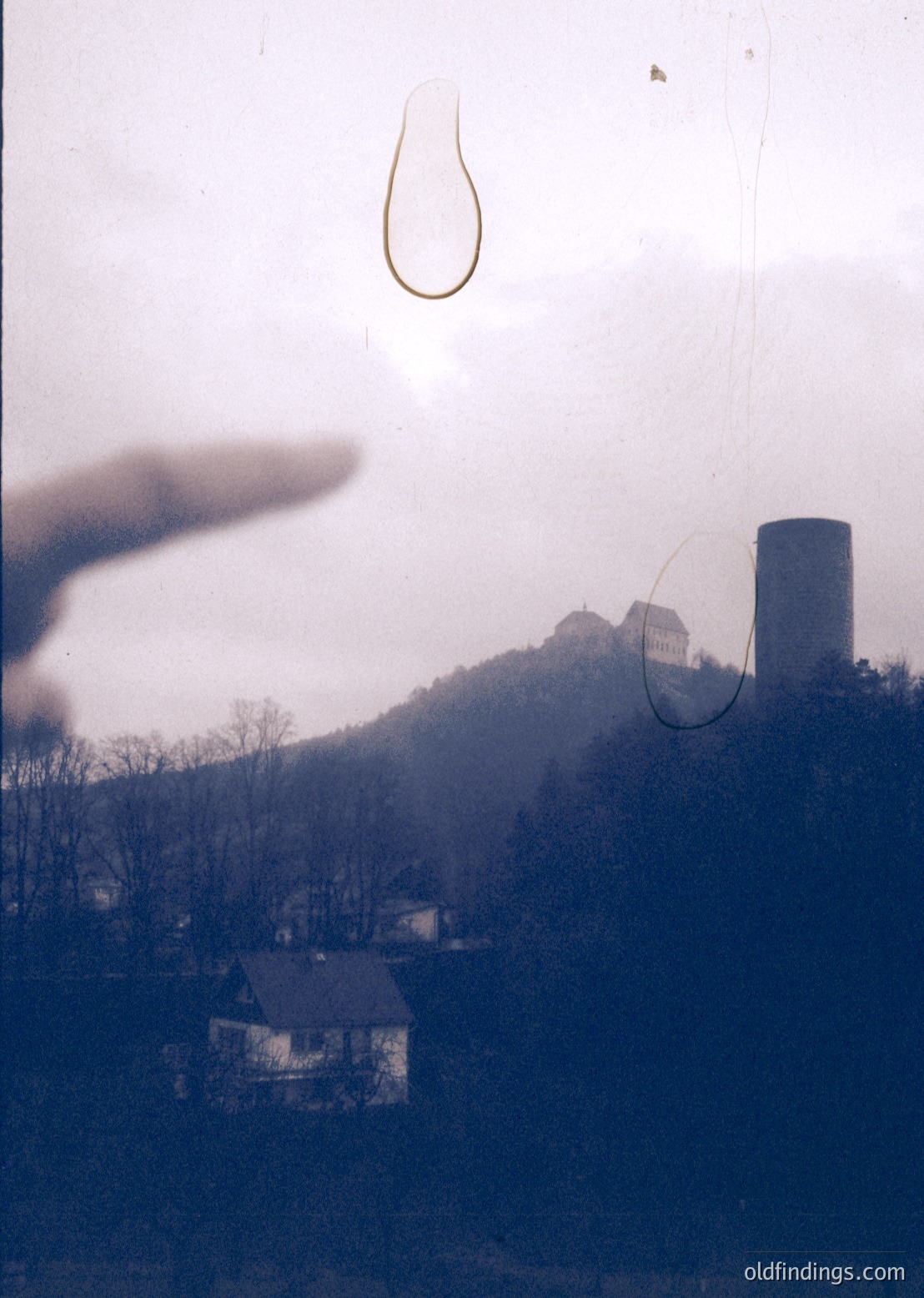 Vintage photograph featuring a blurred, foggy landscape with a lone industrial smokestack and distant castle ruins. Overlaid geometric shapes (teardrop and circle) obscure parts of the scene. Likely mid-20th century European rural setting.