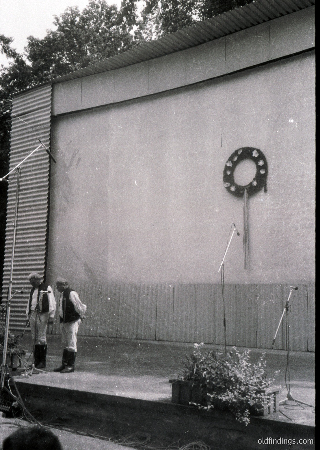 Indoor stage with concrete wall backdrop featuring a wreath emblem. Two men in traditional attire (light-colored robes) stand on a small platform with microphones. Decorative potted plant on stage floor. Likely a cultural or ceremonial event, possibly Soviet-era or Eastern Bloc region.