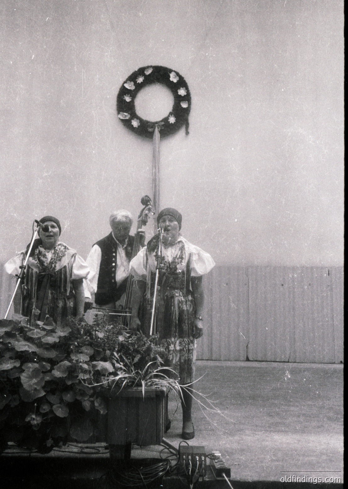 Black-and-white photo of three musicians in traditional folk attire performing on stage, likely Eastern European (1960s–1970s). Centerpiece: wreath with star motif above them. Instruments include accordion, violin, and tambourine. Stage adorned with potted plants and floral arrangements.