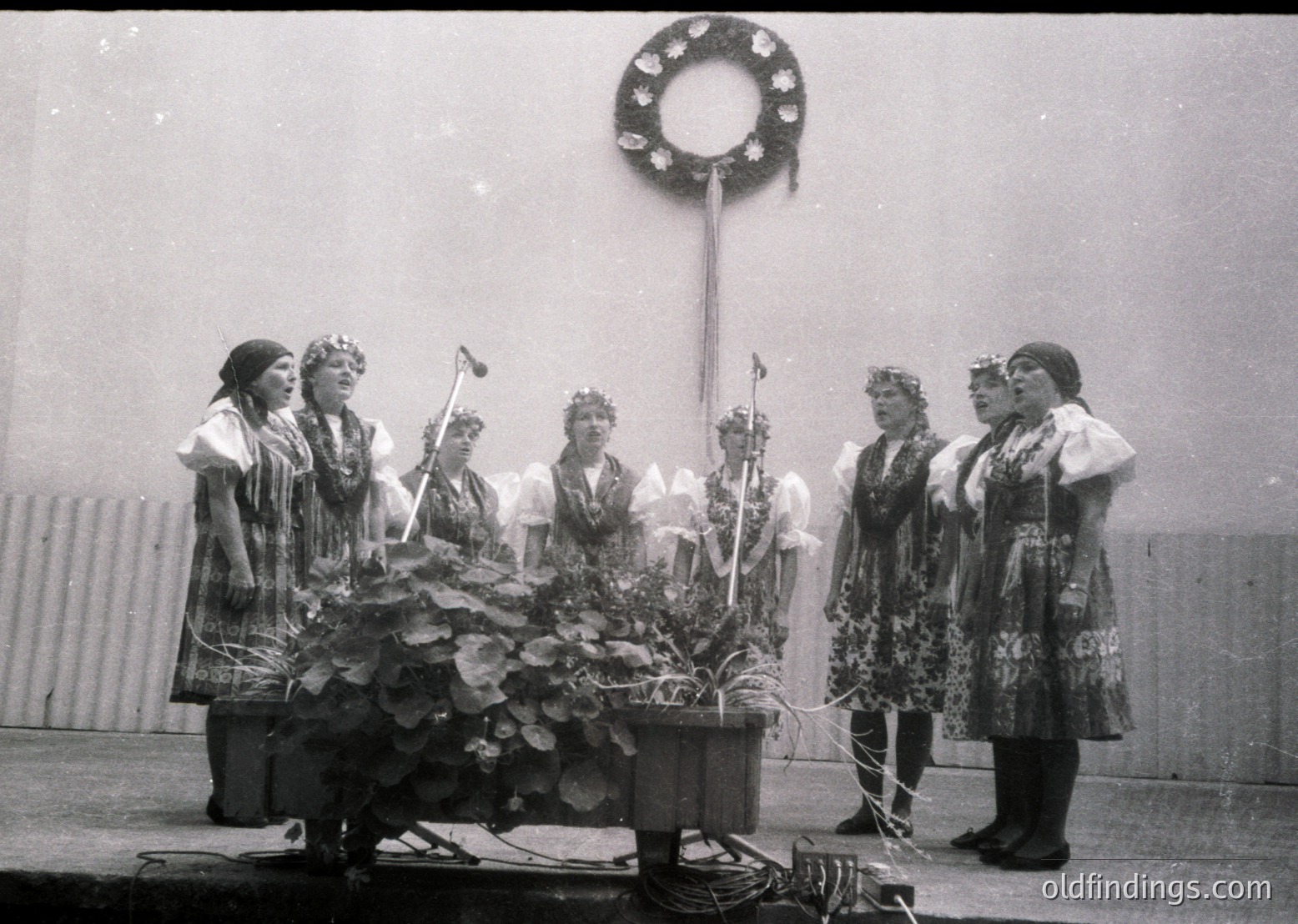 Black-and-white photo of a traditional folk ensemble performing outdoors, likely Eastern European. Seven women in elaborate embroidered costumes with floral headpieces sing or recite around a large floral centerpiece. A wreath hangs overhead, suggesting a ceremonial or festive occasion. Mid-20th century (1950s–1960s) based on attire and grain.