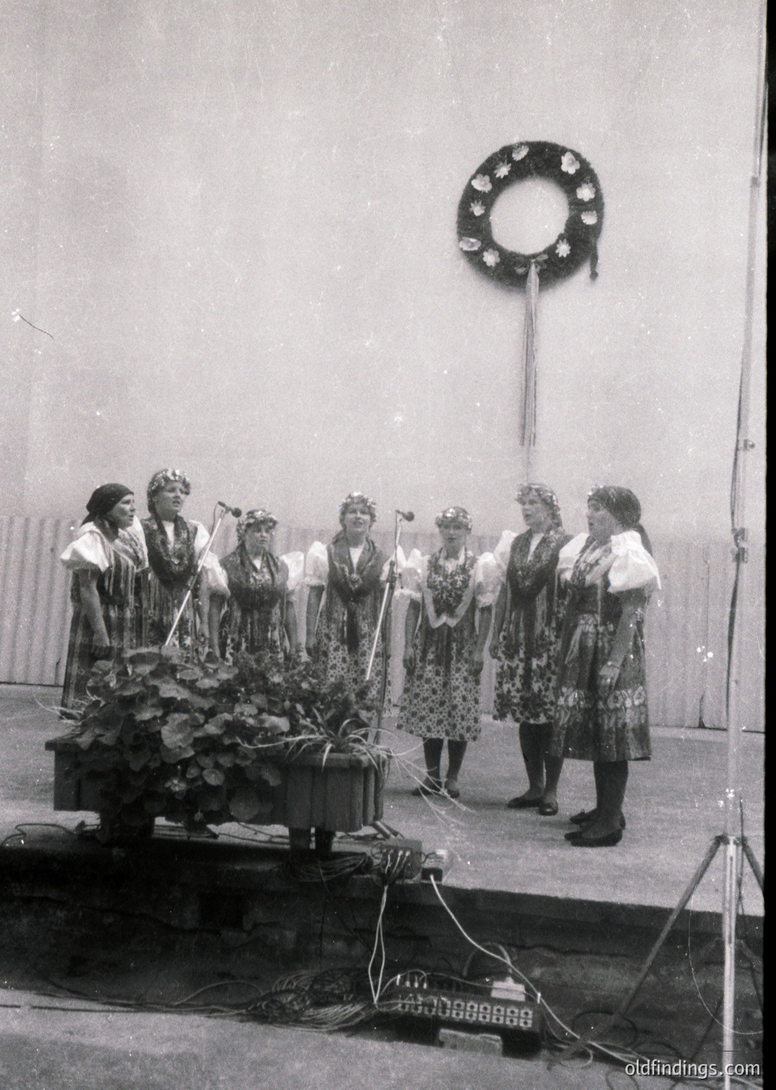 Seven women in traditional folk attire, likely Eastern European, perform on a stage with floral centerpieces. Woven headpieces and embroidered dresses suggest a cultural festival or public celebration. Microphones and a wreath on a pole indicate a formal event, possibly mid-20th century.