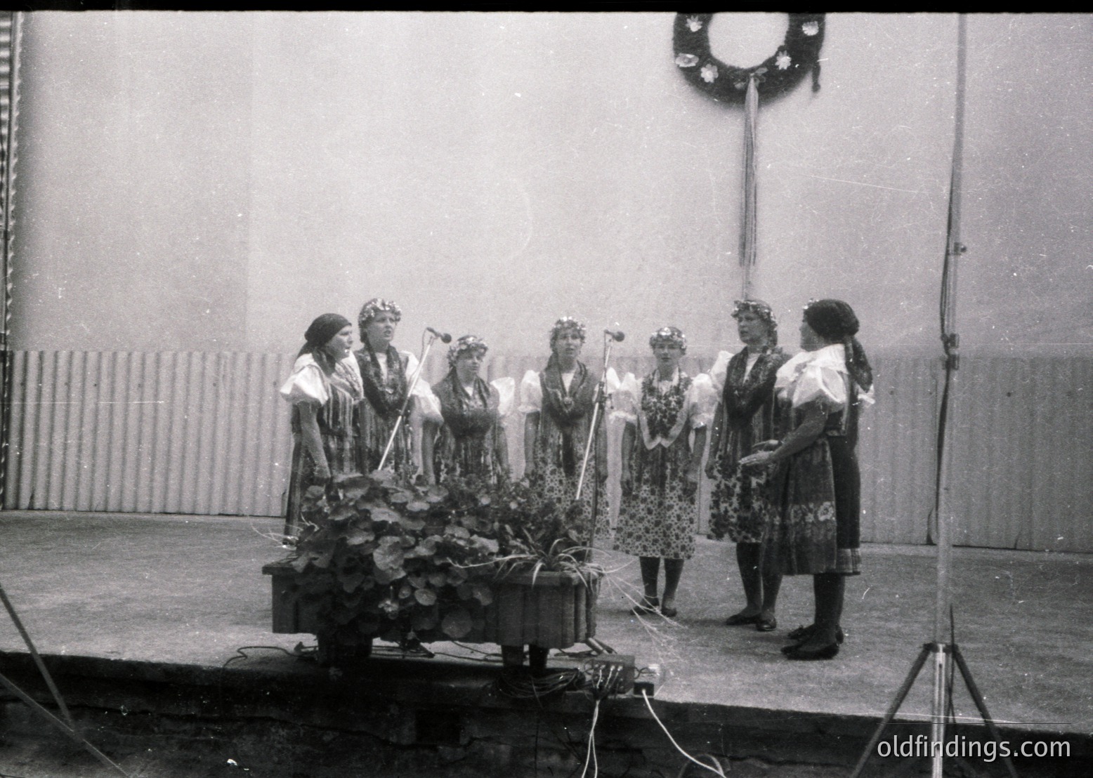 Six women in traditional folk attire perform on a stage, holding floral wreaths and instruments. Mid-20th century Eastern European cultural event, likely Bulgaria or Romania. Stage setup includes a wreath decoration and lighting rigs.