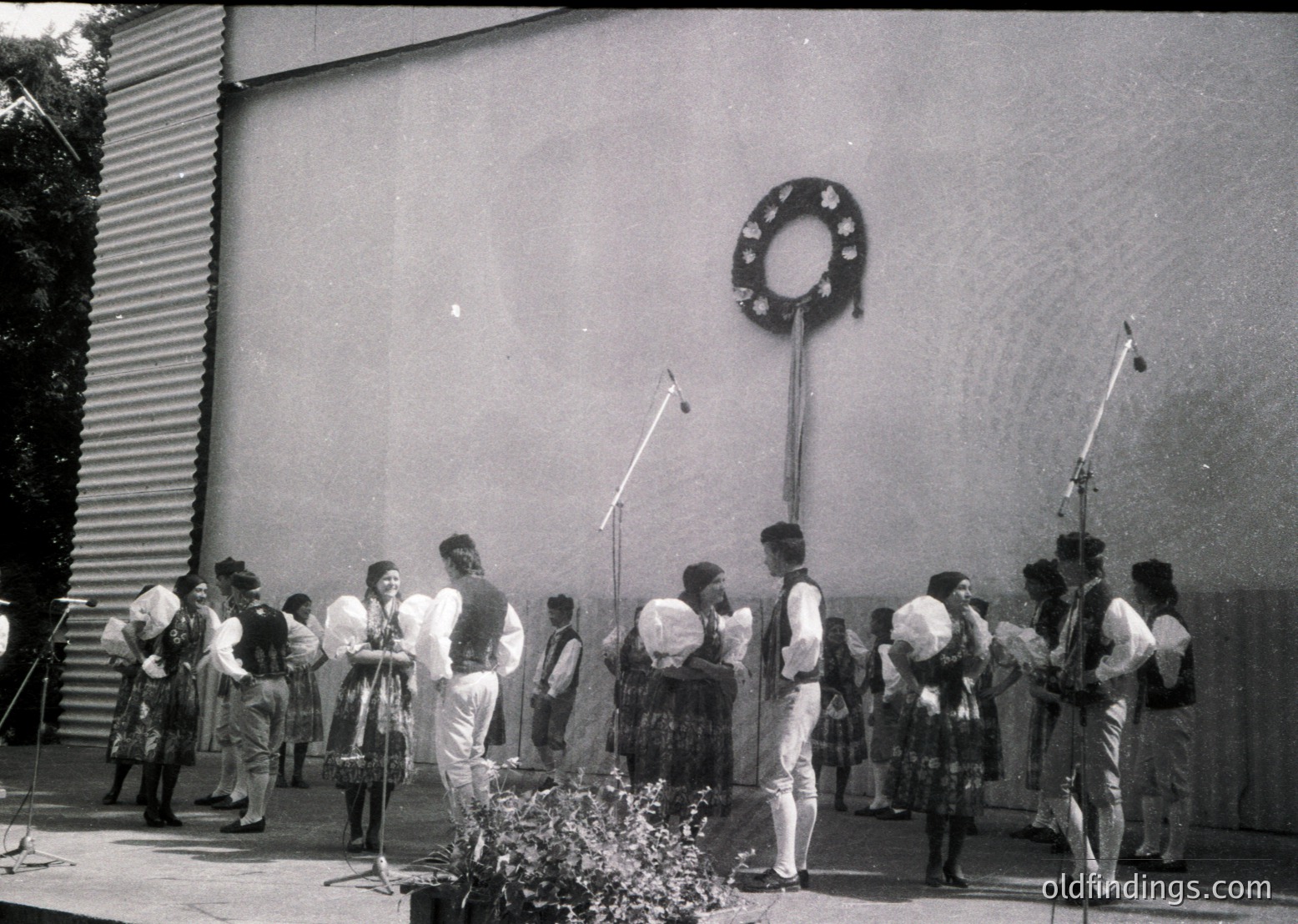 Black-and-white photo of a folk dance ensemble in traditional attire, likely Eastern European, performing outdoors. Men wear white shirts, vests, and knee-length pants with hats; women in embroidered dresses with headscarves. A hammer-and-sickle emblem on a wall suggests Soviet-era Bulgaria (). Ideal for historical research or cultural documentation.