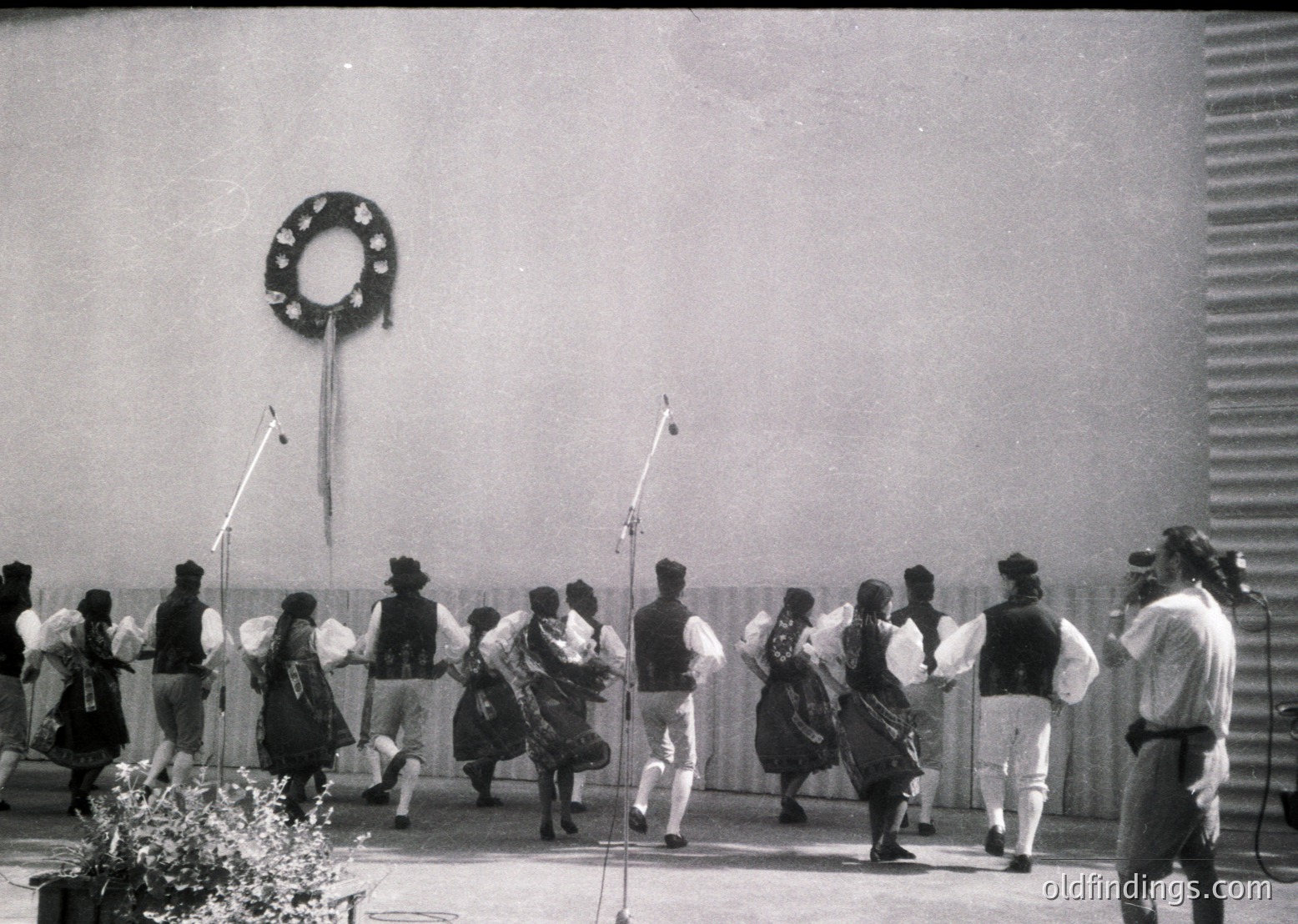 Traditional folk dancers in regional attire perform outdoors, likely in Eastern Europe. Men wear vests, knee-length pants, and wide-brimmed hats; women in layered dresses with aprons. A microphone stand suggests live music accompaniment. Decorative wreath on wall hints at cultural celebration. Mid-20th century (1950s–1970s) folk festival.