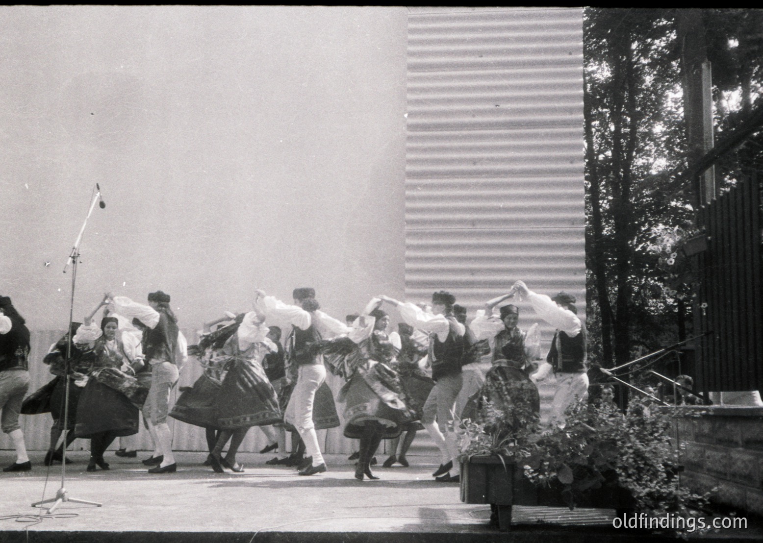 Traditional folk dancers in elaborate costumes perform outdoors, likely in a 1960s–1970s Eastern European setting. Men wear white trousers with sashes, women feature layered skirts and embroidered blouses. Microphone stand and modernist building with horizontal slats in background.