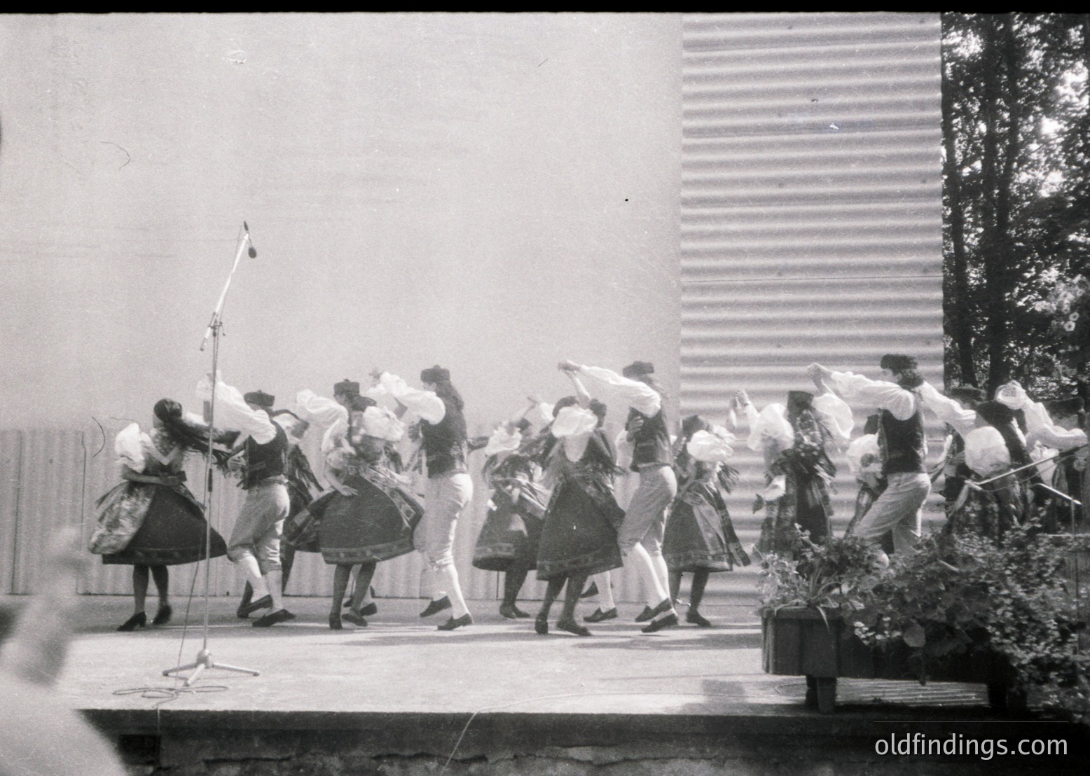 Group of women performing traditional folk dance in coordinated attire—long skirts, blouses, and headscarves—under a microphone stand. Mid-20th century outdoor setting with industrial-style backdrop. Likely Eastern European cultural event.