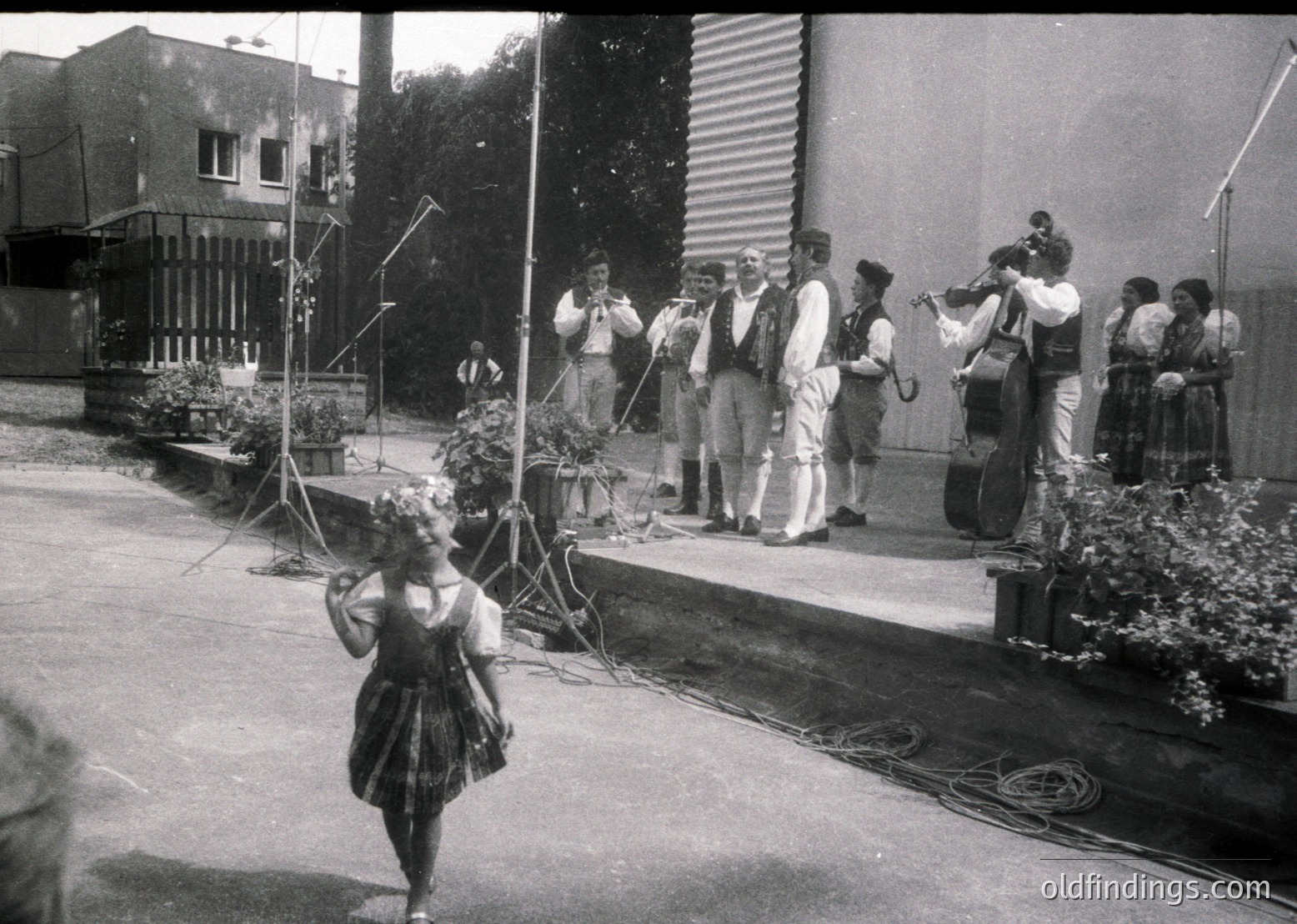 Black-and-white street performance featuring a young dancer in traditional skirt and headpiece, accompanied by a live folk orchestra on a raised platform. Instruments include accordions, violins, and a cello. Mid-20th century European outdoor cultural event, likely or .