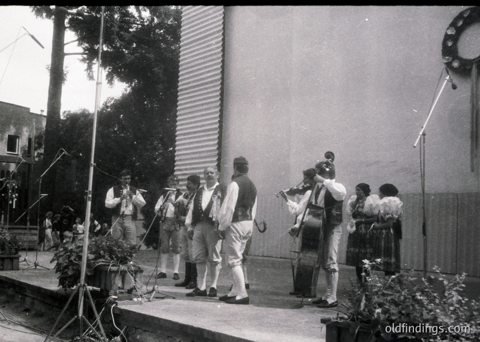 Black-and-white street performance featuring a traditional folk ensemble in 1960s-70s attire: accordion, violin, cello, and percussion. Stage setup includes microphones and a simple backdrop of corrugated metal. Audience stands casually behind barriers. Urban setting with visible trees and residential buildings.