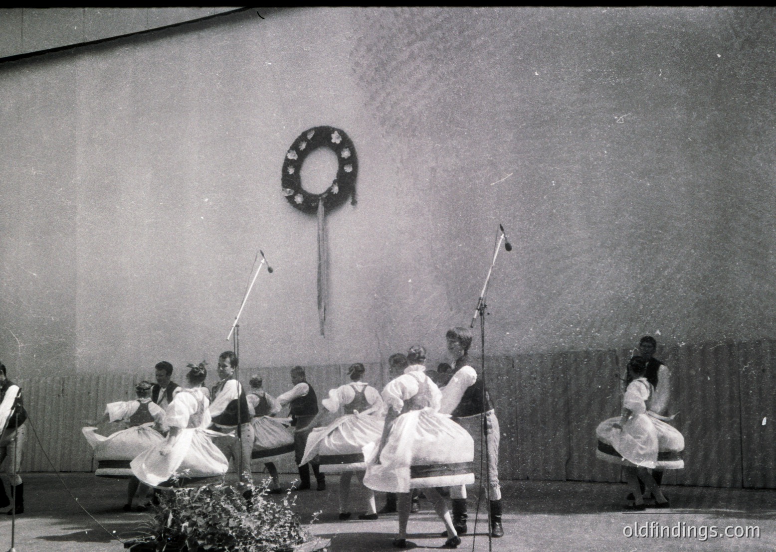 Black-and-white stage performance featuring women in traditional folk costumes, likely Eastern European, performing a circular dance. Stage backdrop includes a wreath emblem and curtain. Mid-20th century (1950s–1960s) cultural event.