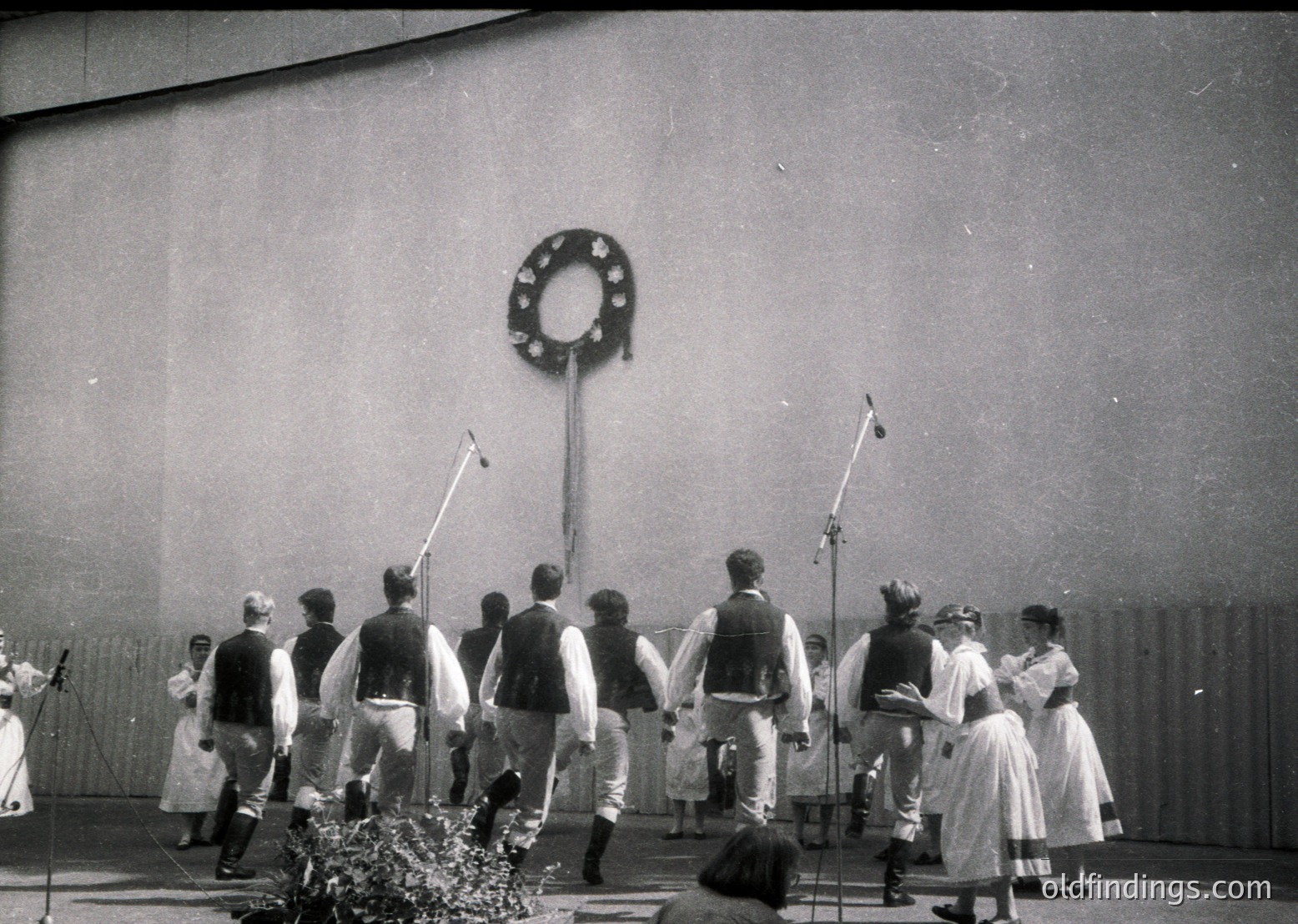 Black-and-white photo of a traditional folk dance performance, likely Eastern European, featuring men in vests, knee-length pants, and headgear, paired with women in long dresses and floral headbands. A wreath with a hammer-and-sickle emblem hangs on a wall, suggesting a mid-20th century Soviet-era setting. Microphones and musical instruments indicate a staged event.
