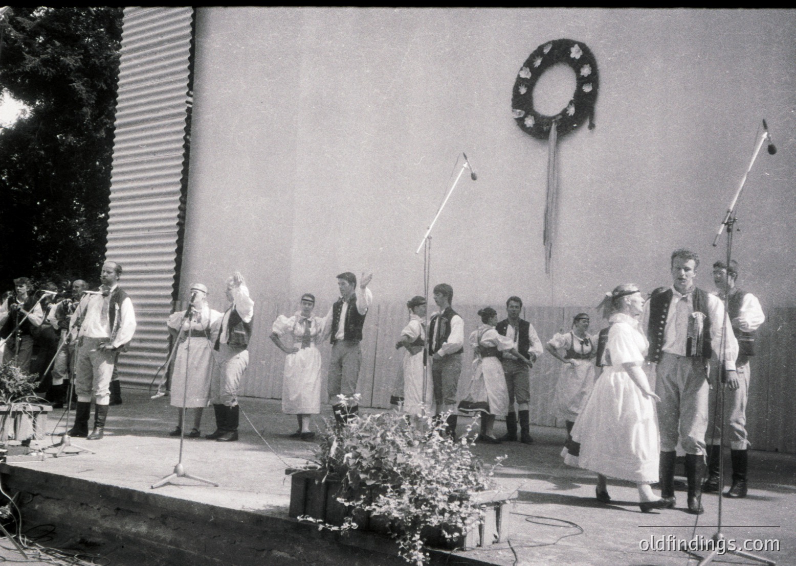 Group performing traditional folk dance on stage, likely Eastern European . Men in vests, women in embroidered dresses and headscarves. Stage backdrop features wreath and industrial-style wall. Microphones and floral centerpiece enhance cultural event ambiance.