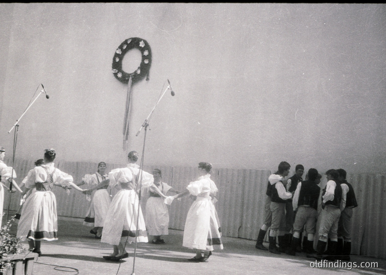 Black-and-white photo of a traditional folk dance performance, likely Eastern European, featuring women in white embroidered dresses and men in dark trousers with vests. The circular formation and decorative wall hook suggest a cultural or festive event, possibly mid-20th century. Stage lighting and microphone stands indicate a formal or organized setting.