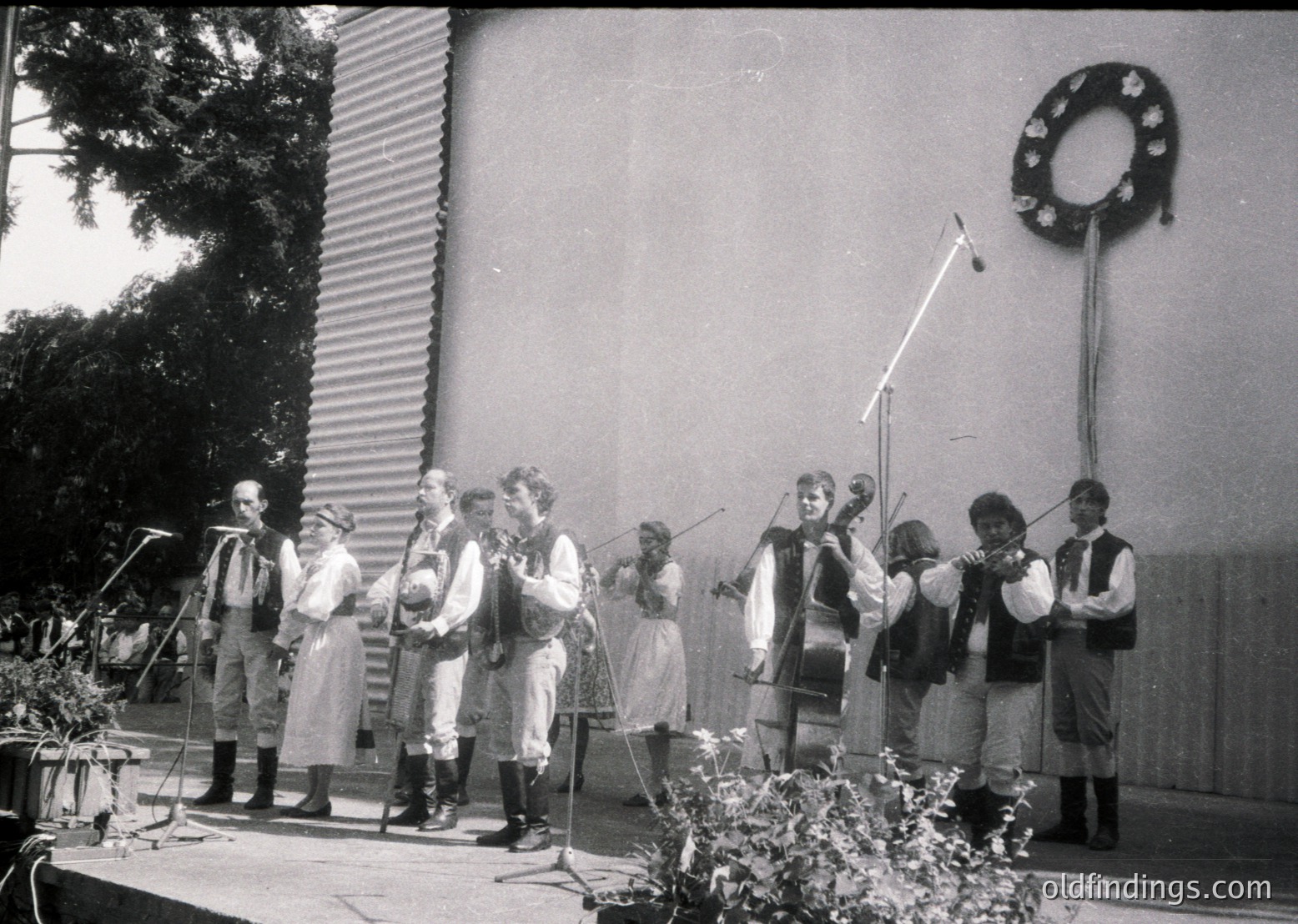 Traditional folk ensemble performing outdoors in 1960s Eastern European attire—men in embroidered vests and women in long skirts, playing string instruments. Decorative wreath on wall suggests cultural festival or public event.