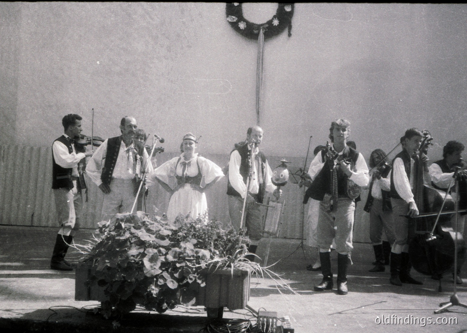 Traditional folk ensemble performing in traditional attire, likely Eastern European (1960s-70s). Seven musicians—four playing string instruments (violins, accordion), two on percussion—surround a central singer in folk dress. Decorative floral arrangement at center suggests a festive or cultural event. Indoor setting with simple stage backdrop.