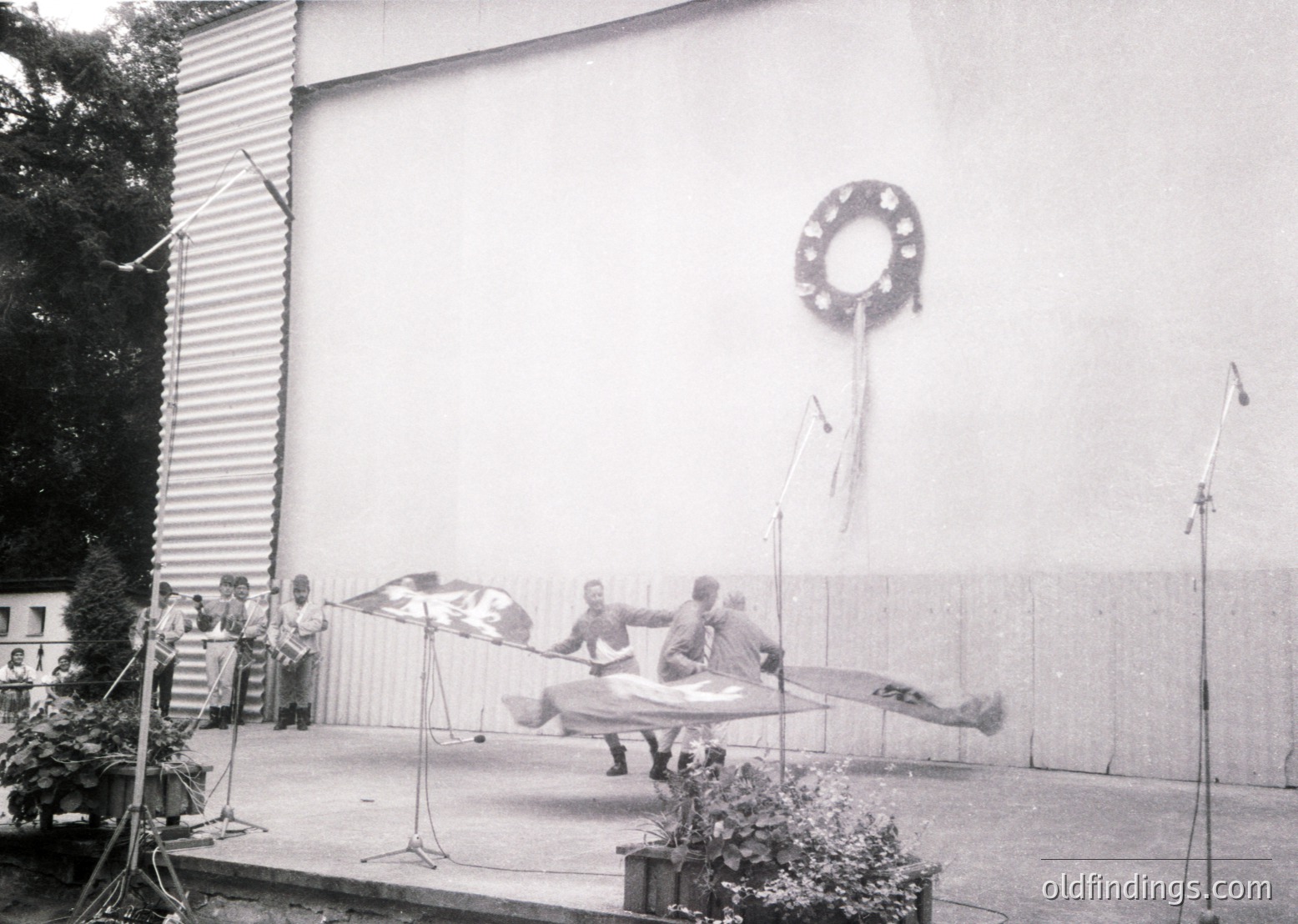 Black-and-white shot of a 1960s-70s stage performance featuring a trapeze act. Four performers mid-air on a suspended trapeze bar, with a backdrop of a plain wall and decorative wreath. Audience seated on tiered bleachers. Industrial-style lighting and microphone stands visible.