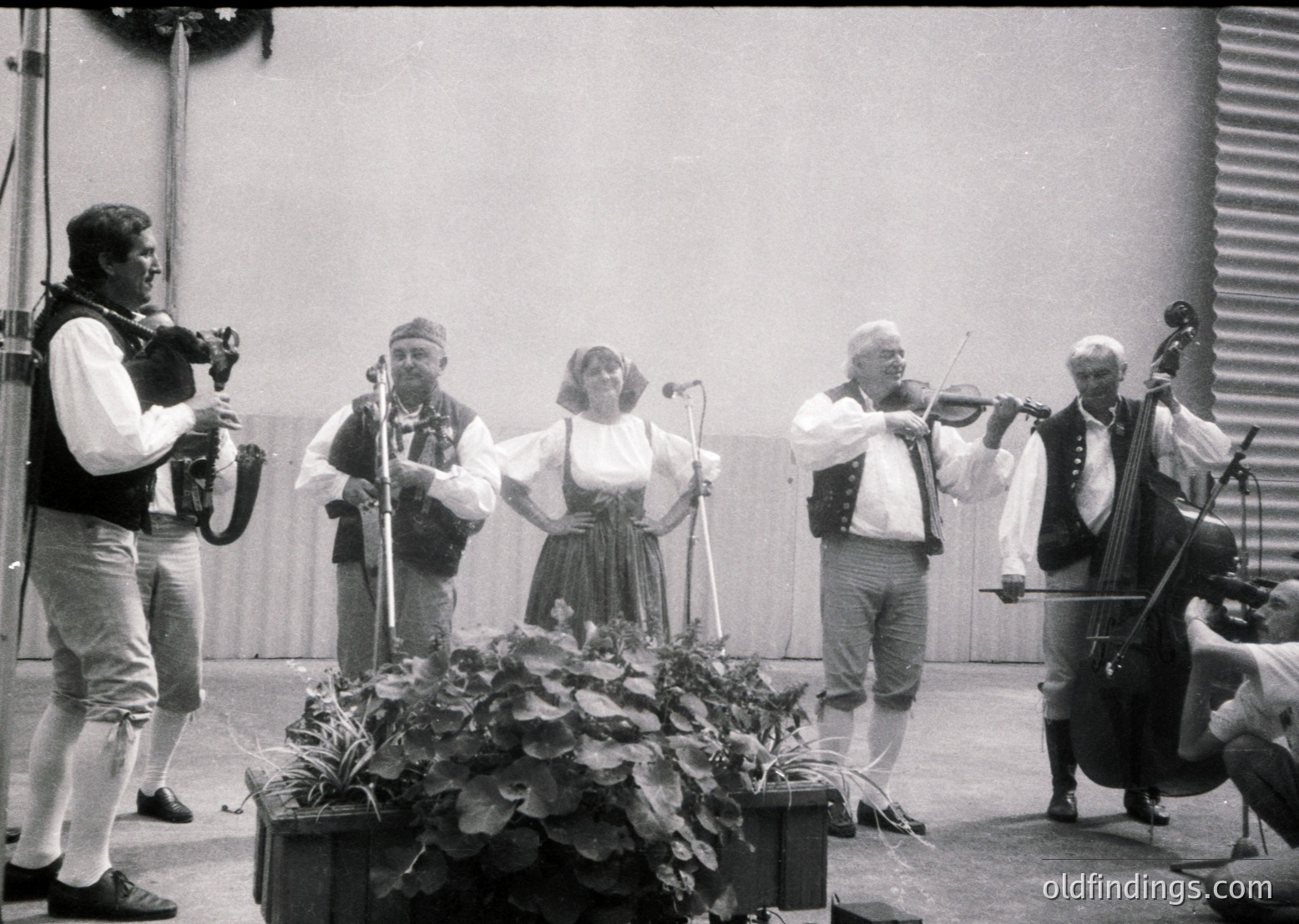 Five musicians perform outdoors in folk attire, likely Eastern European . Violinists, a cellist, accordionist, and vocalist stand on a wooden platform with floral centerpiece. Industrial backdrop suggests a rural festival or cultural event.