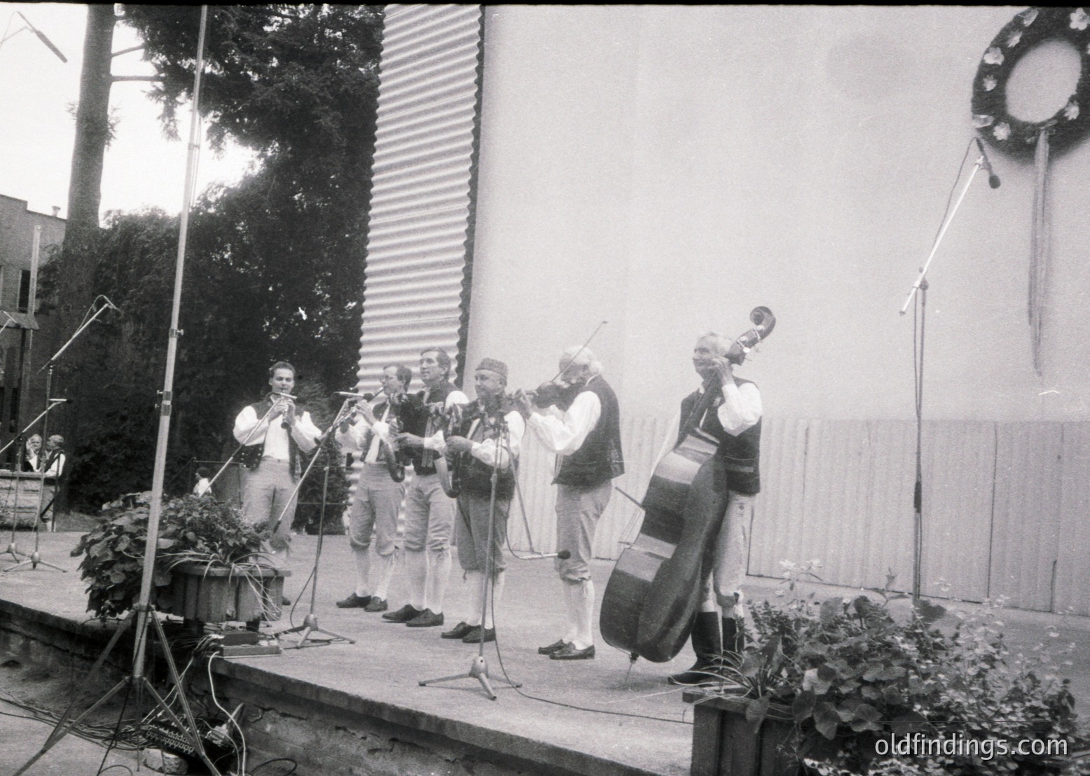 Six musicians perform outdoors on a stage with a corrugated metal backdrop. Instruments include violins, a cello, and a saxophone. Decorative potted plants flank the stage. Likely a 1960s–1980s Eastern Bloc cultural event.