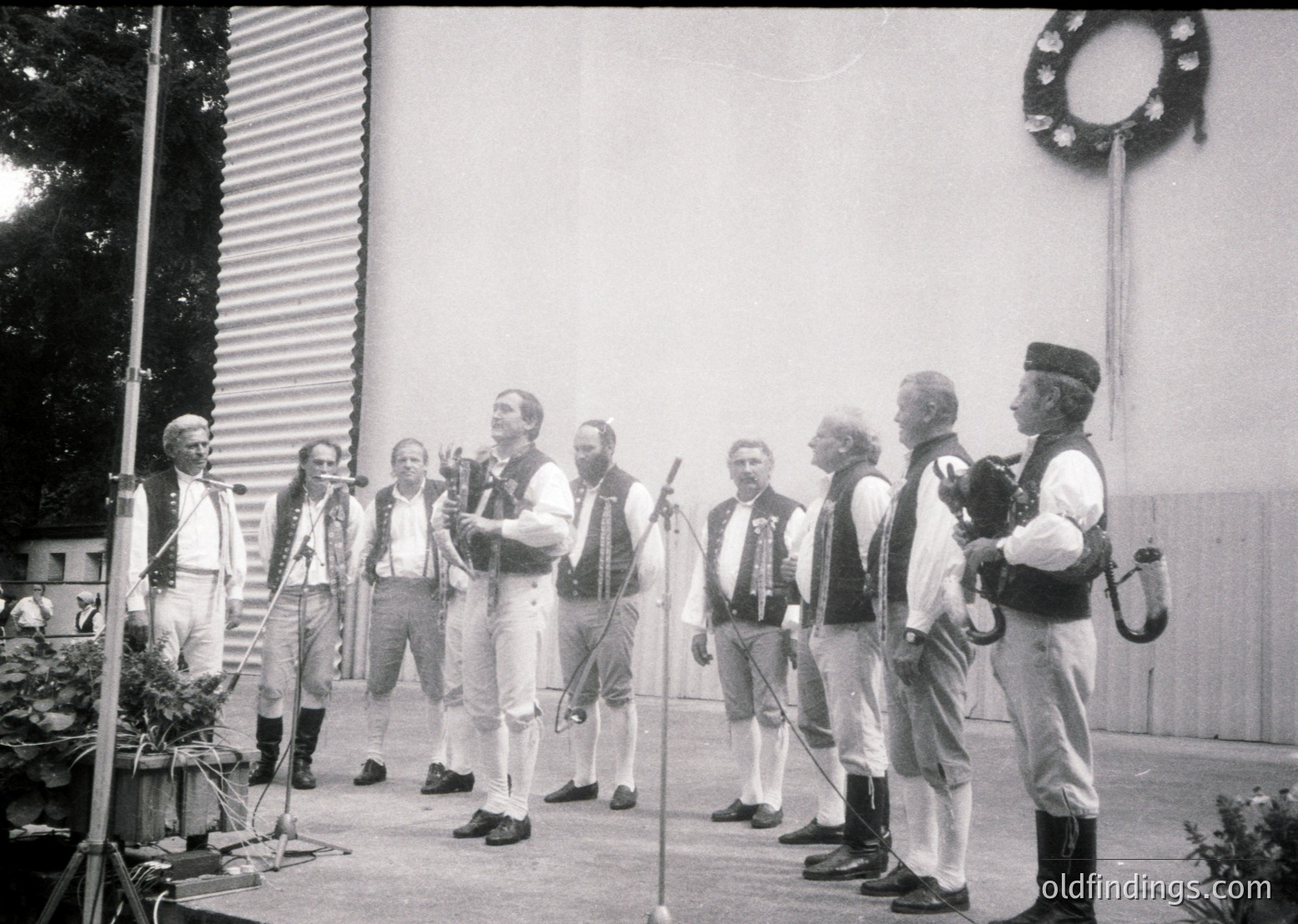 Eight men in traditional folk attire perform outdoors, likely Eastern European given attire and wreath. Central figure holds microphone, others play stringed instruments. Industrial-style backdrop suggests 1960s–1980s era.