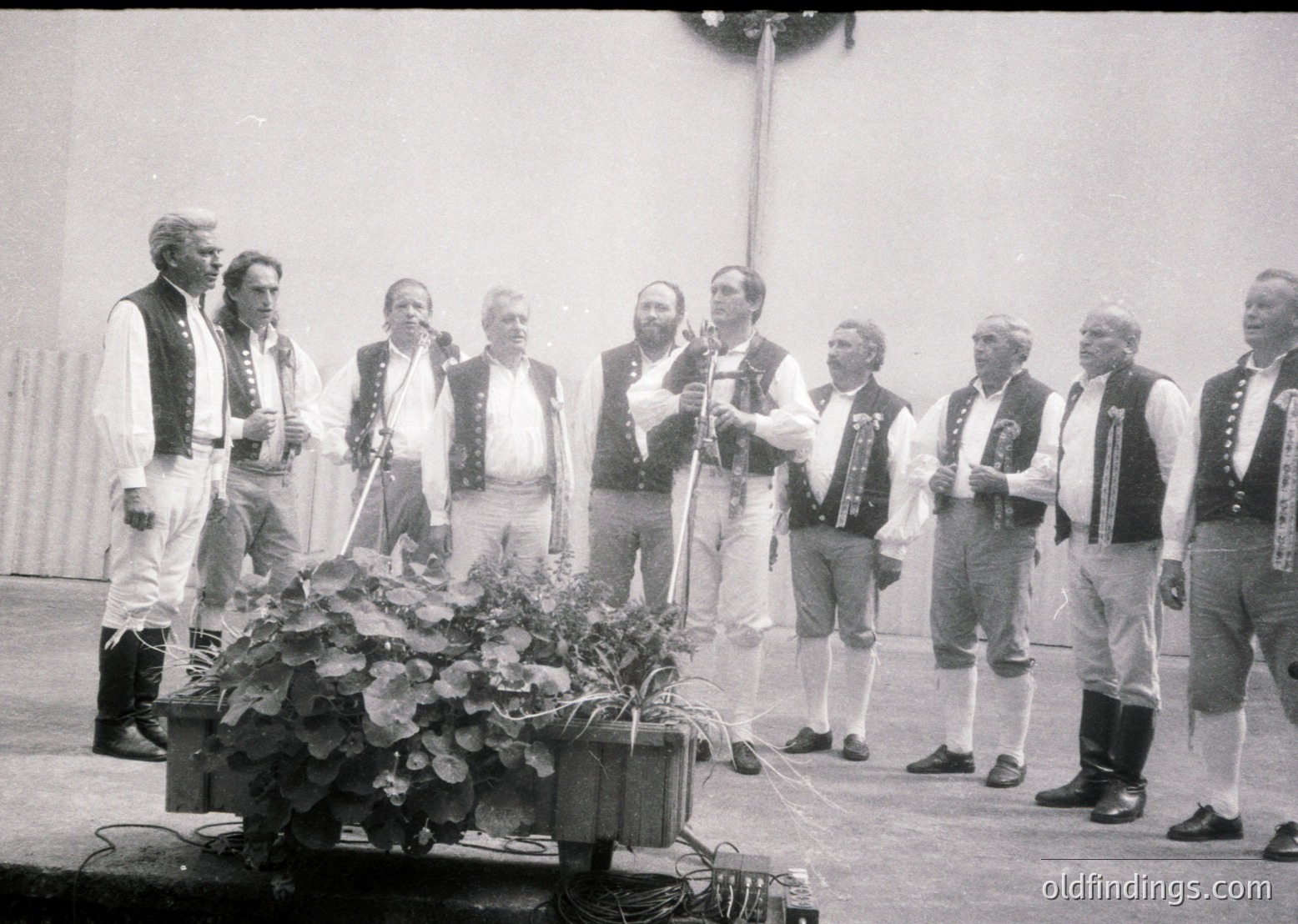 Men in traditional Balkan folk attire perform outdoors, likely in a 1960s–1980s Eastern European setting. Group stands in a semi-circle around a floral centerpiece, suggesting a cultural festival or folk music event. Distinctive embroidered vests, knee-high socks, and headwear highlight regional heritage.