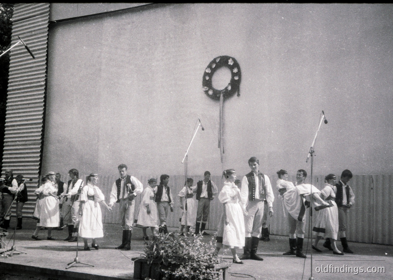A black-and-white group performance featuring 14 individuals in traditional folk attire, likely Eastern European, performing outdoors. Men wear vests and neckerchiefs; women in long dresses with aprons. Microphones and a wreath on a pole suggest a staged event, possibly a festival or cultural celebration. Mid-20th century (1950s–1960s) based on clothing and photography style.