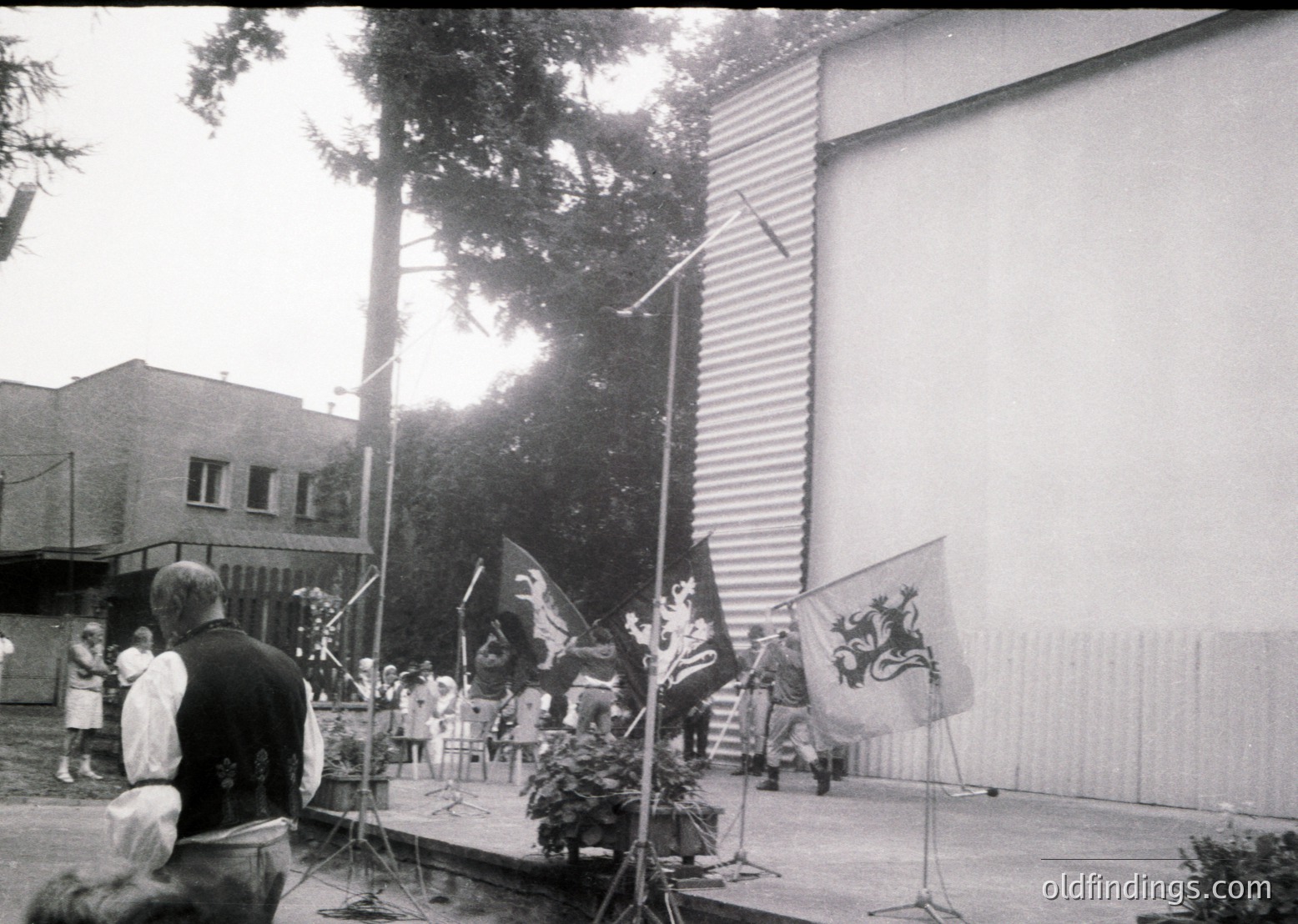 Black-and-white shot of a formal outdoor ceremony, likely 1960s–1980s Eastern Bloc. Stage features floral centerpiece, three flags with heraldic crests, and a podium. Attendees in formal attire (suits, dresses) stand or sit in rows. Industrial-style building and trees frame the scene. Potential historical research or Cold War-era documentation.