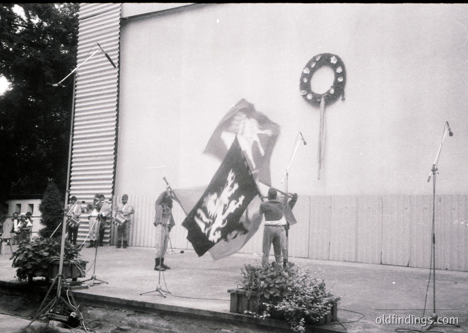 Black-and-white photo of a public ceremony featuring a large banner with a hammer-and-sickle emblem and text in Cyrillic. Stage setup includes microphones, a wreath on a pole, and a floral arrangement. Attendees in formal attire, likely mid-20th century Eastern Bloc.