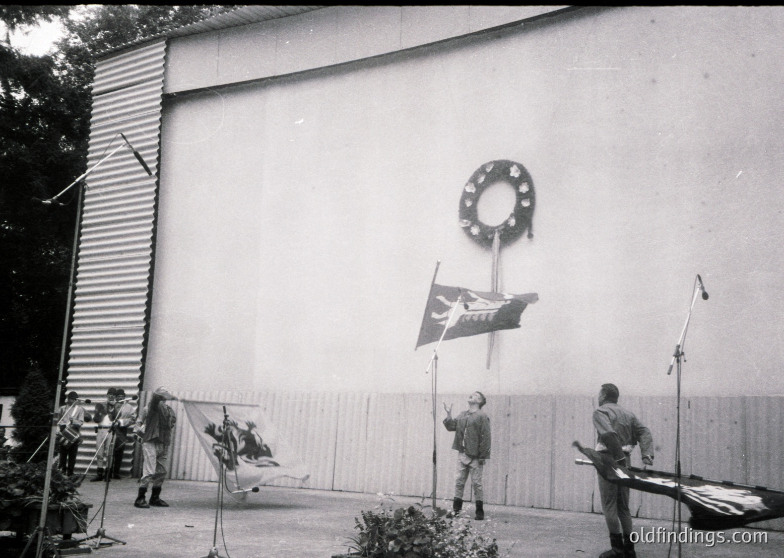 Mid-20th century formal ceremony at a modernist building with clean lines. A wreath and flagpole with a flag bearing a hammer-and-sickle emblem dominate the center. Six men in suits and caps stand in a semi-circle, some holding microphones. Microphone stands and a draped backdrop suggest an official event.