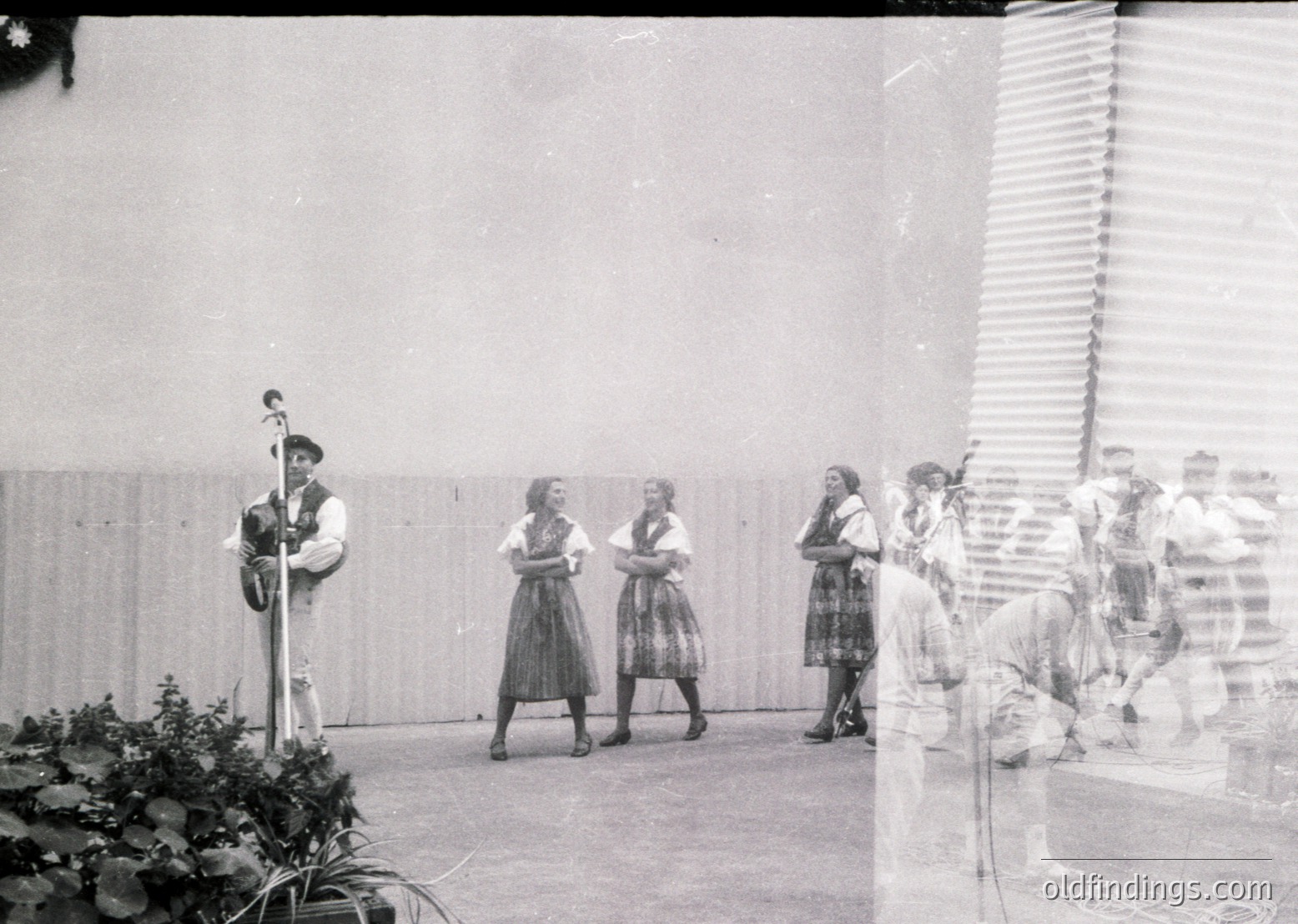 Black-and-white group performance featuring a man playing a stringed instrument (likely a bandura) and women in traditional folk attire with pleated skirts and embroidered blouses, likely Eastern European. Indoor setting with plain walls and potted plants.