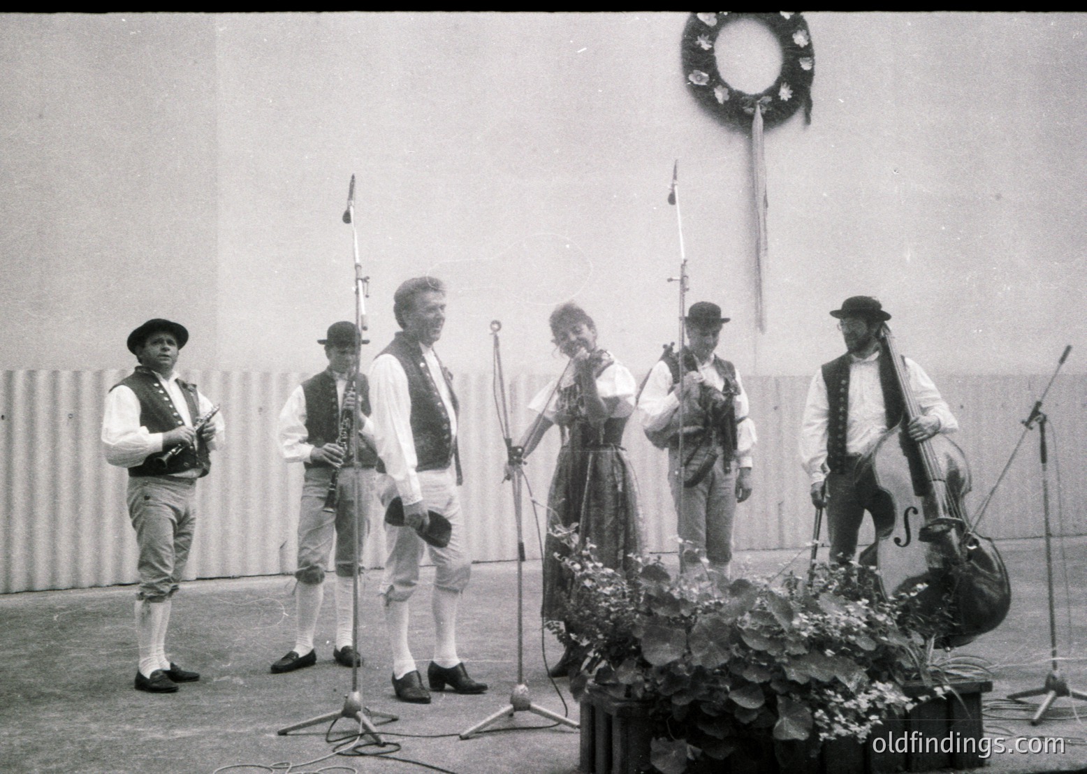 Traditional folk ensemble performing outdoors, likely Eastern European . Five musicians in folk attire—fiddles, accordion, flute, violin, and cello—positioned around a decorated table with floral centerpiece. Industrial backdrop suggests a public event or festival.