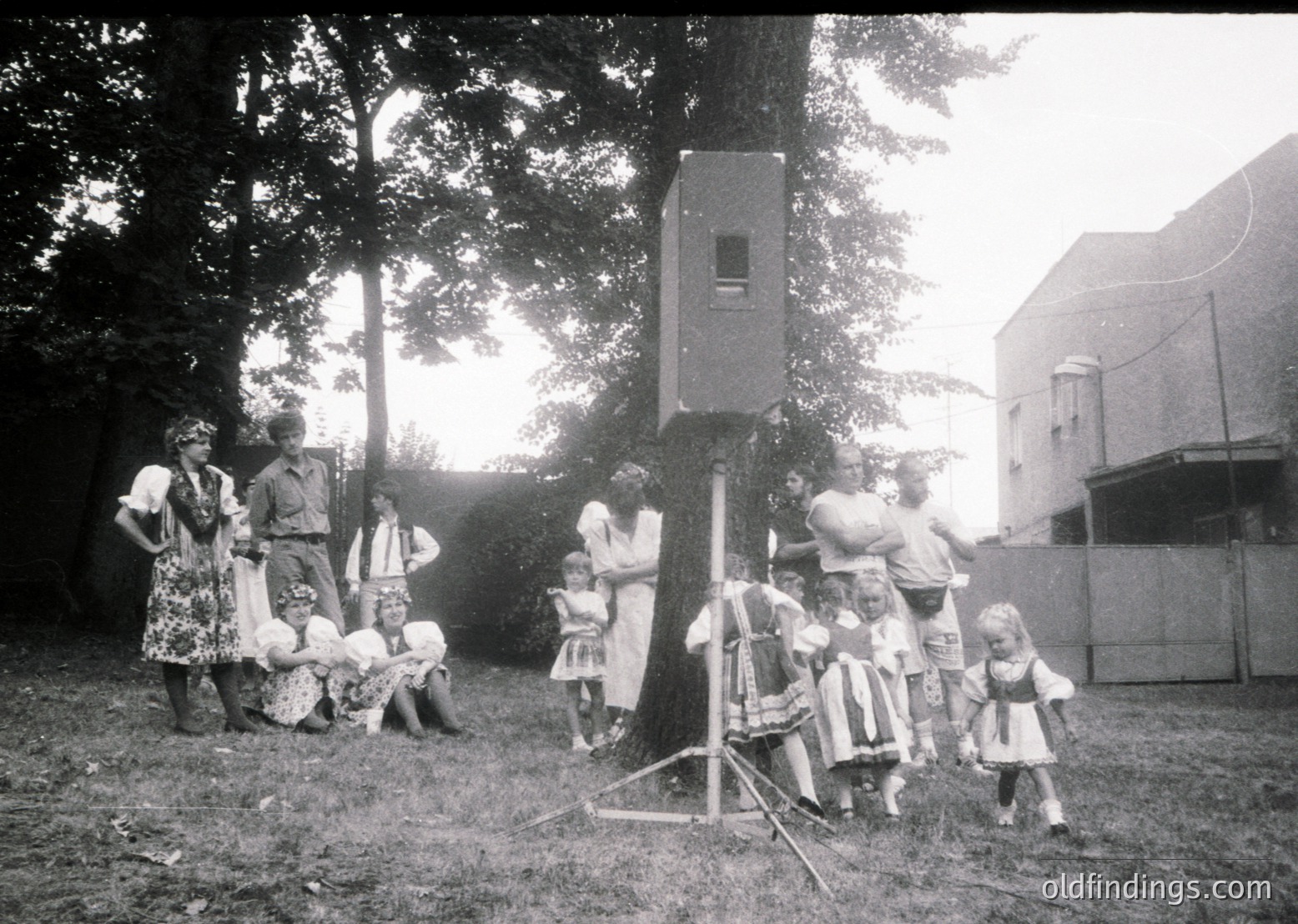 Vintage black-and-white photo of a mid-20th century outdoor gathering, likely 1950s–1960s. Group of adults and children pose around a vintage telephone booth-style booth with a speaker, suggesting a community event or public announcement system. Attire includes floral dresses, suits, and headscarves, reflecting mid-century Western European or North American style. Lush greenery and a tree frame the scene, indicating a park or backyard setting.
