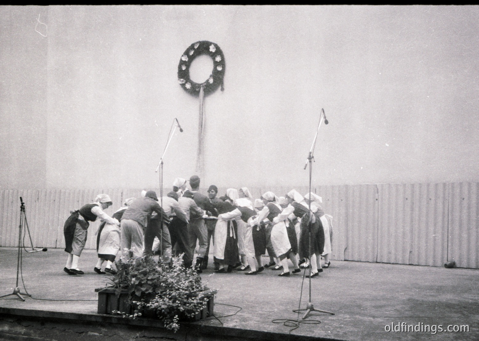 Group of women in traditional folk attire performing a circle dance, likely a Bulgarian *horo*, under a decorative metal ring prop. Stage setup includes microphones and floral centerpiece. Mid-20th century (1950s–1970s) cultural event.